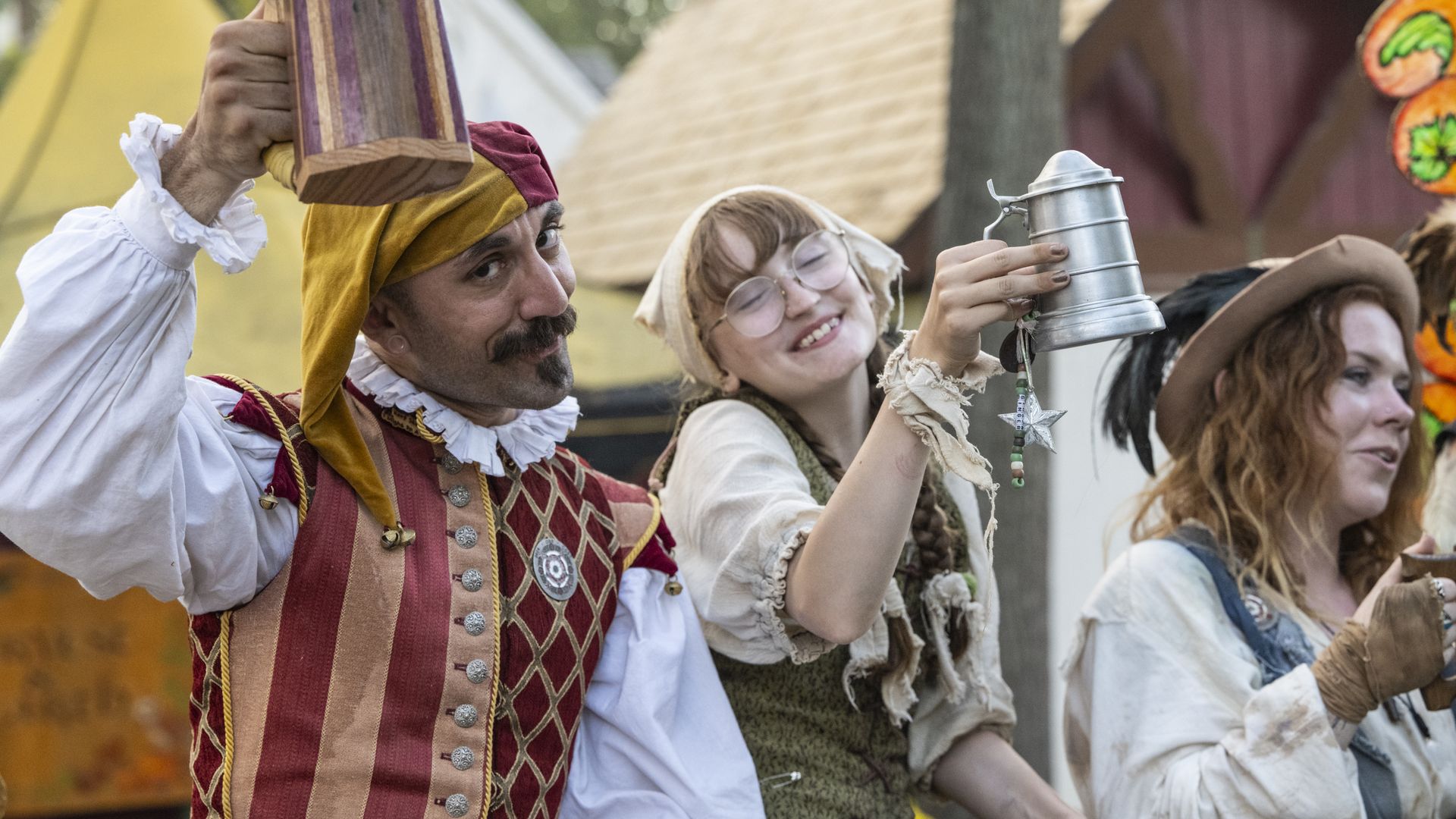 Three people in Renaissance-style costumes at a fair, raising drinking mugs, smiling, with colorful tents in the background.