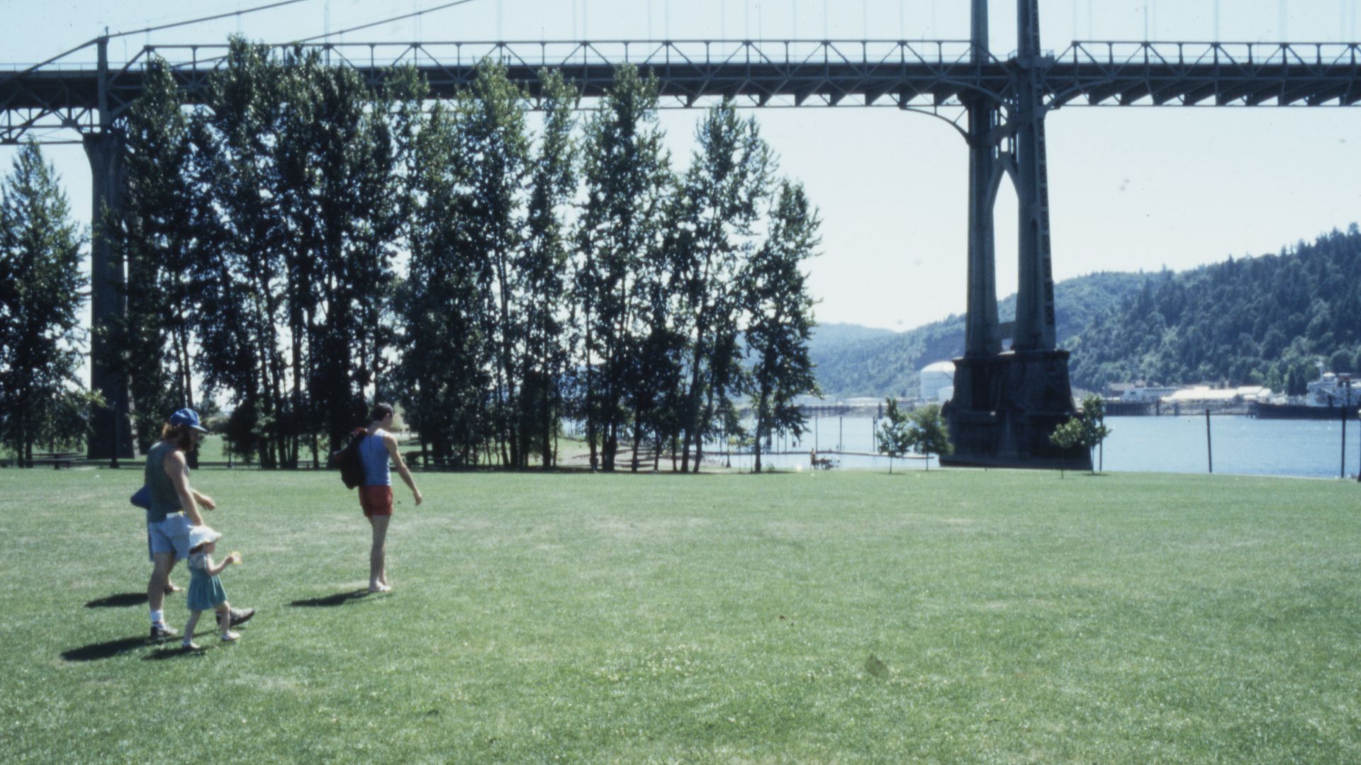 People walk on a grassy riverside park beneath a tall steel arch bridge; trees line the bank, with water and hills in the background.