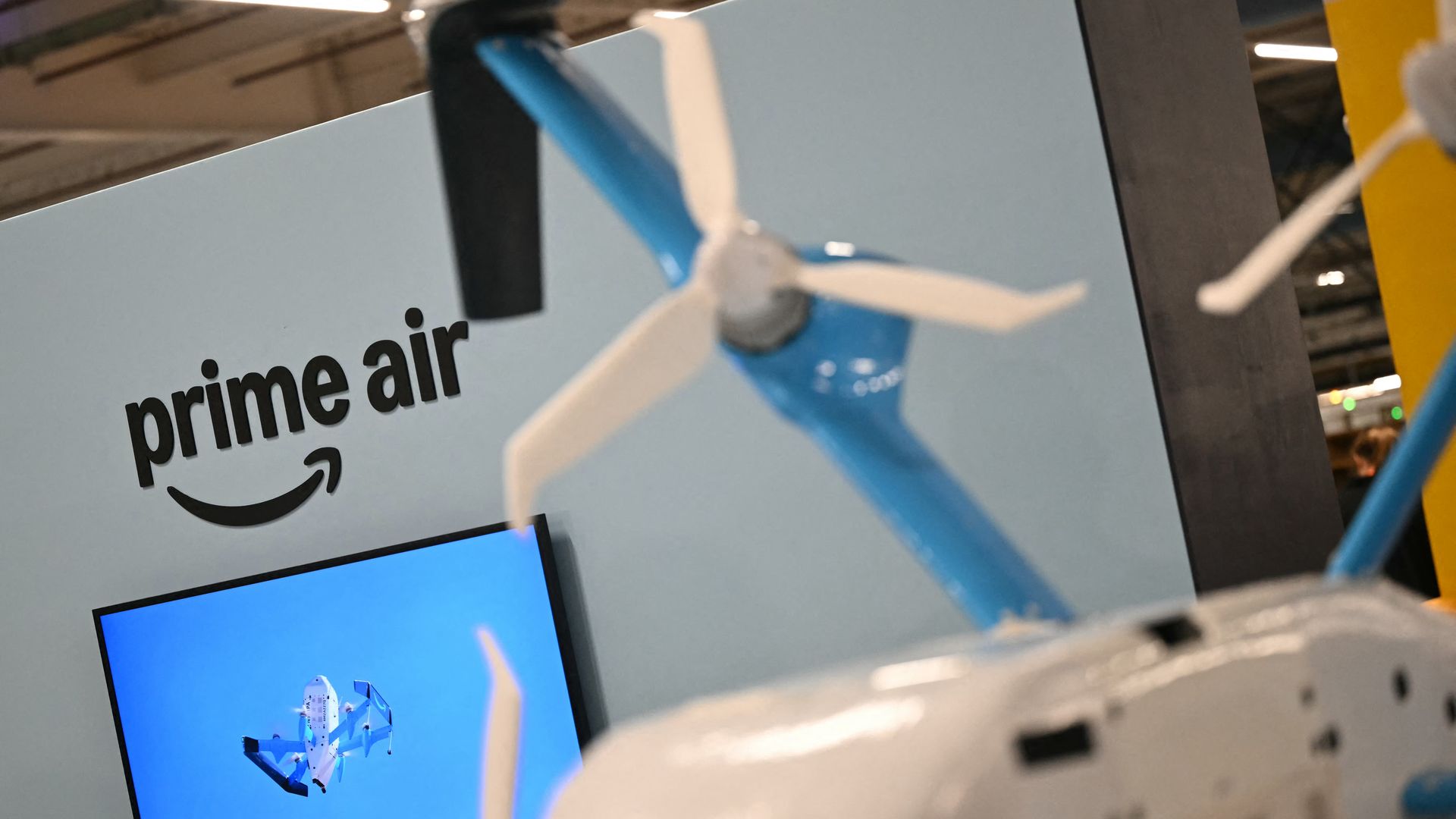 Exhibit panel reads "prime air" with Amazon logo; a blue drone propeller crosses the foreground. A blue monitor shows a drone image; blurred background includes a yellow pillar.