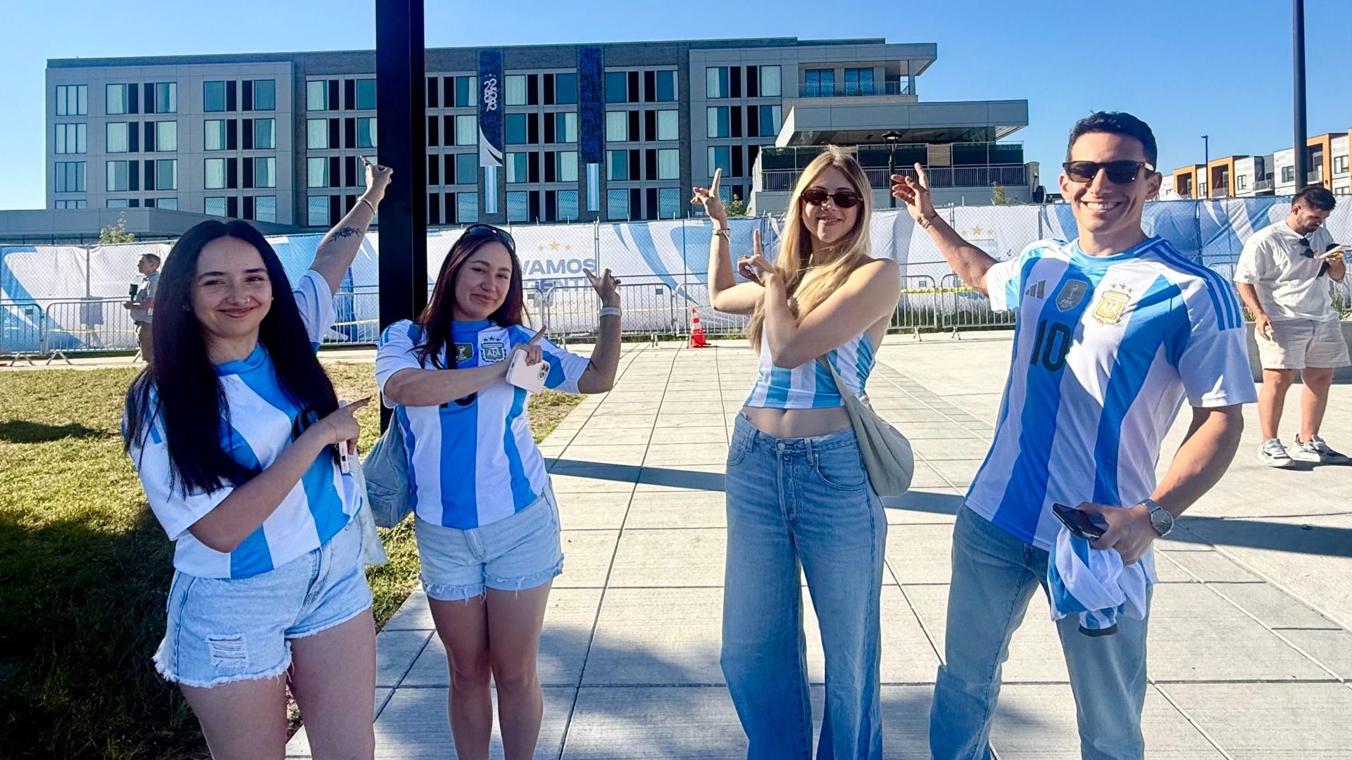Four people wearing Argentina blue-and-white soccer jerseys pose outdoors on a sunny day, smiling and pointing. A modern building, fence, and banner are in the background.