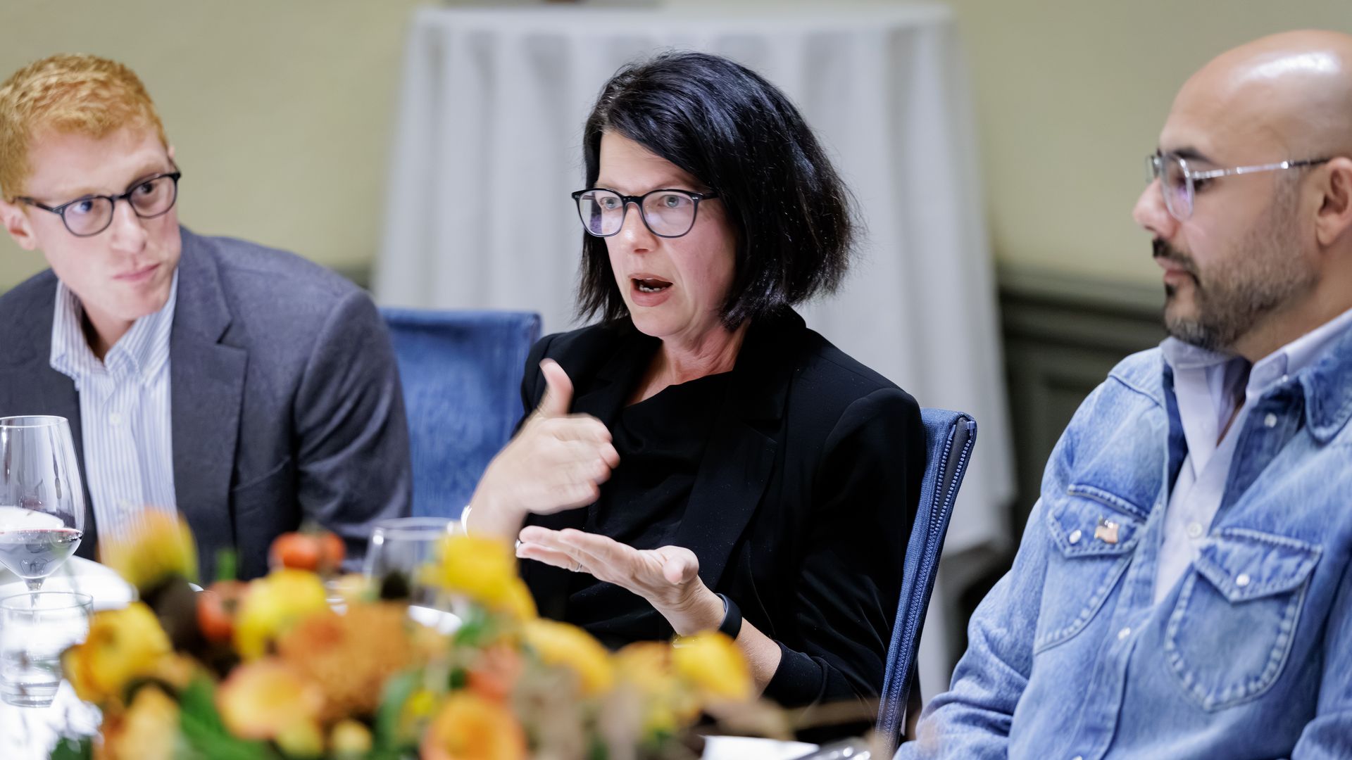 Three people sit at a conference table. Center woman with black hair and glasses speaks, gesturing. Left man in a suit watches; right man in a denim jacket listens. Fruit centerpiece foreground.