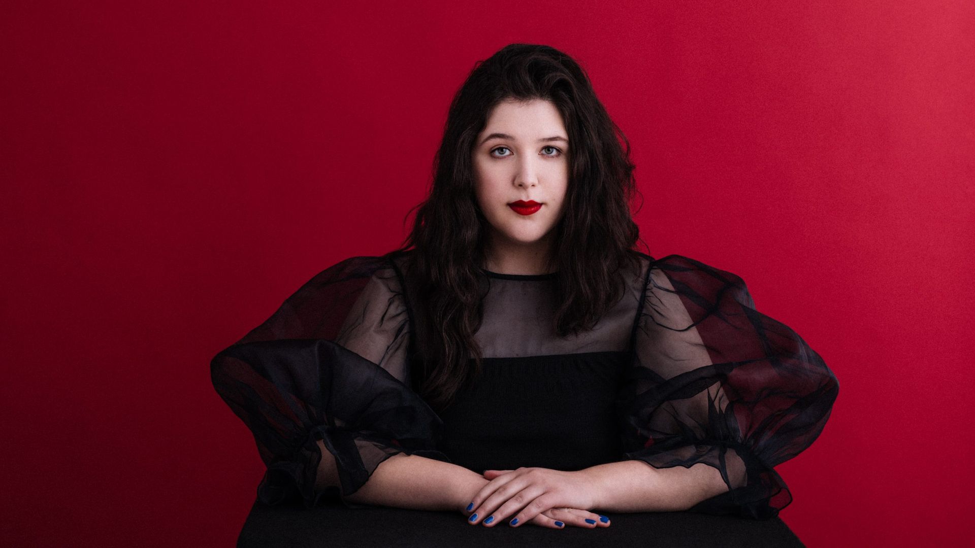 Lucy Dacus poses for a portrait in a black dress in front of a crimson red background.