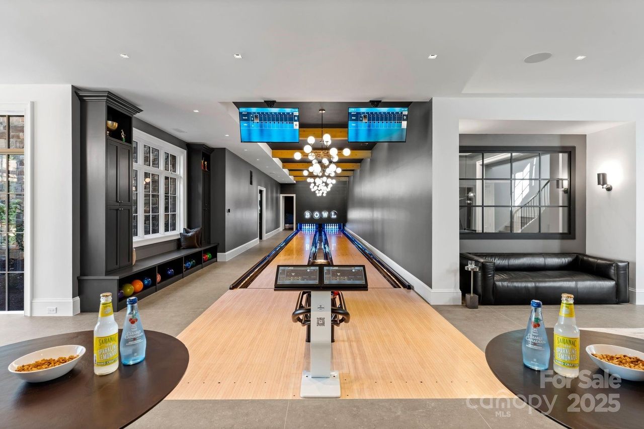 Modern indoor bowling alley with two lanes, dark gray walls, wooden ceiling beams, black leather couch, two round tables with drinks and snacks, and large windows letting in natural light.