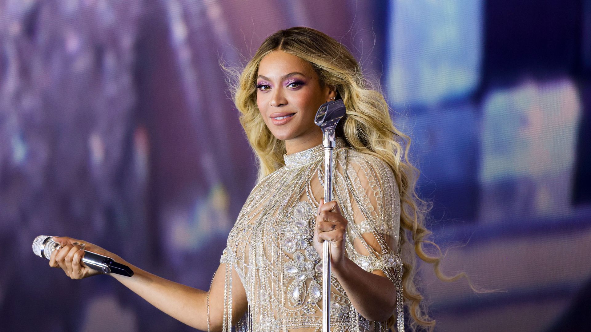 Beyoncé with long blonde hair wearing a silver beaded gown with fringe, holding a microphone and standing at a silver mic stand on stage with a purple backdrop.