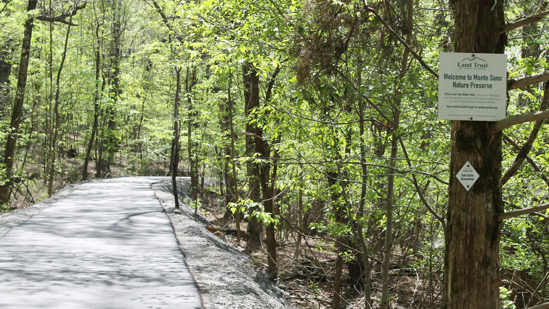 Sunlit forest path winds through green trees; on the right, a tree bears a sign reading "Welcome to Monte Sano Nature Preserve," with a small diamond sign beneath.