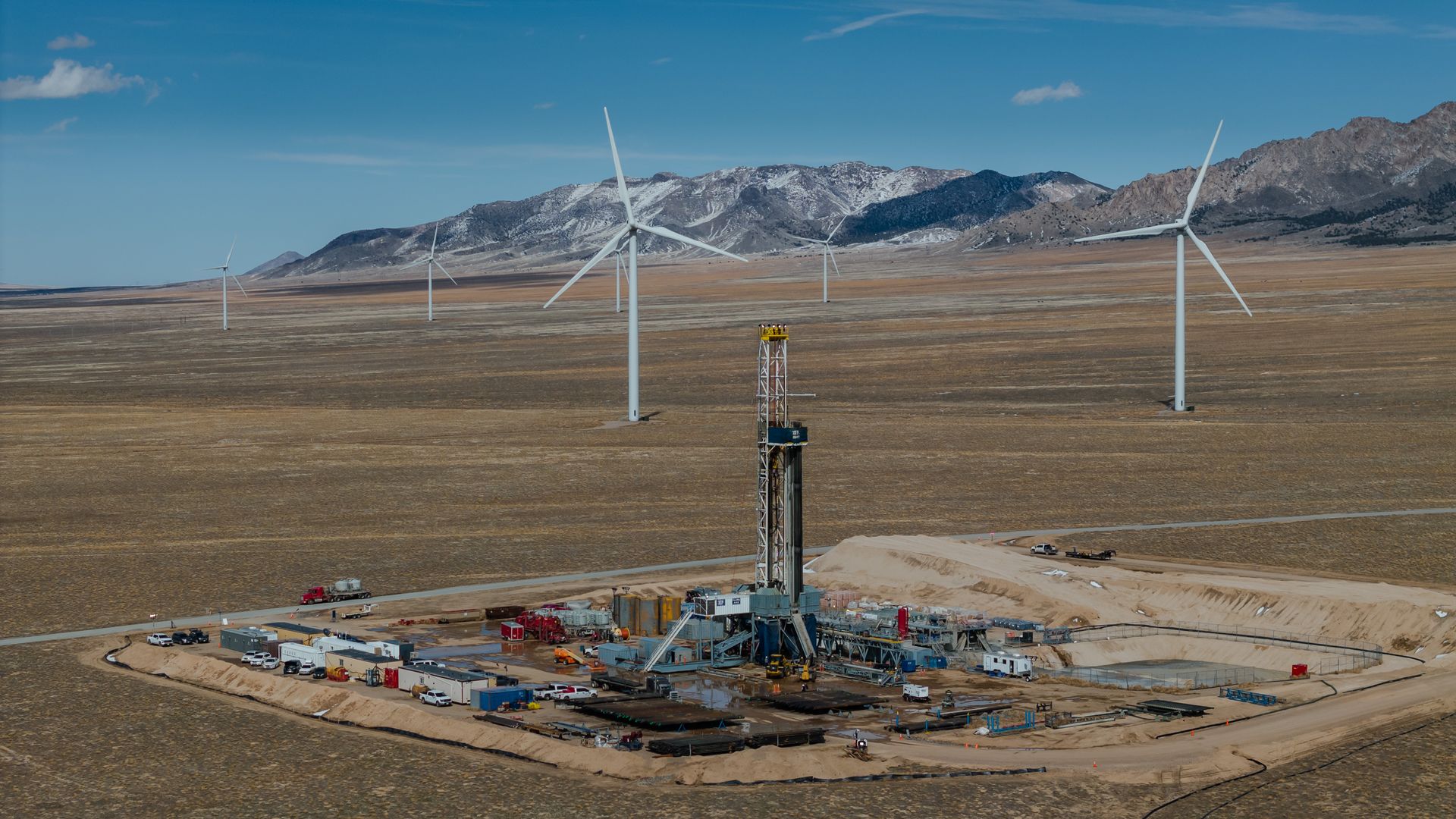Desert plain with several white wind turbines across brown ground, distant snow-capped mountains beneath a blue sky; a construction site and equipment are visible in the foreground.