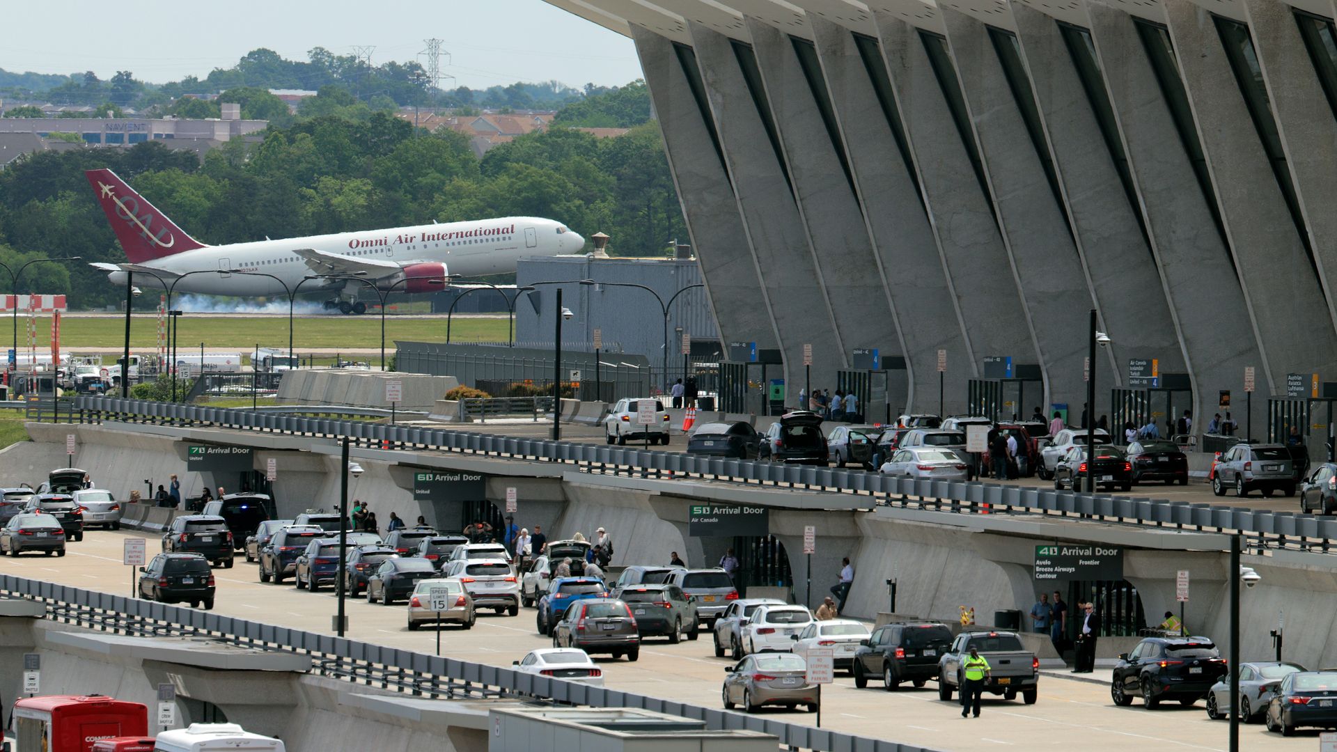Busy airport terminal with multiple cars lining up at arrival doors beneath a large modern concrete building, while an Omni Air International plane touches down in the background.