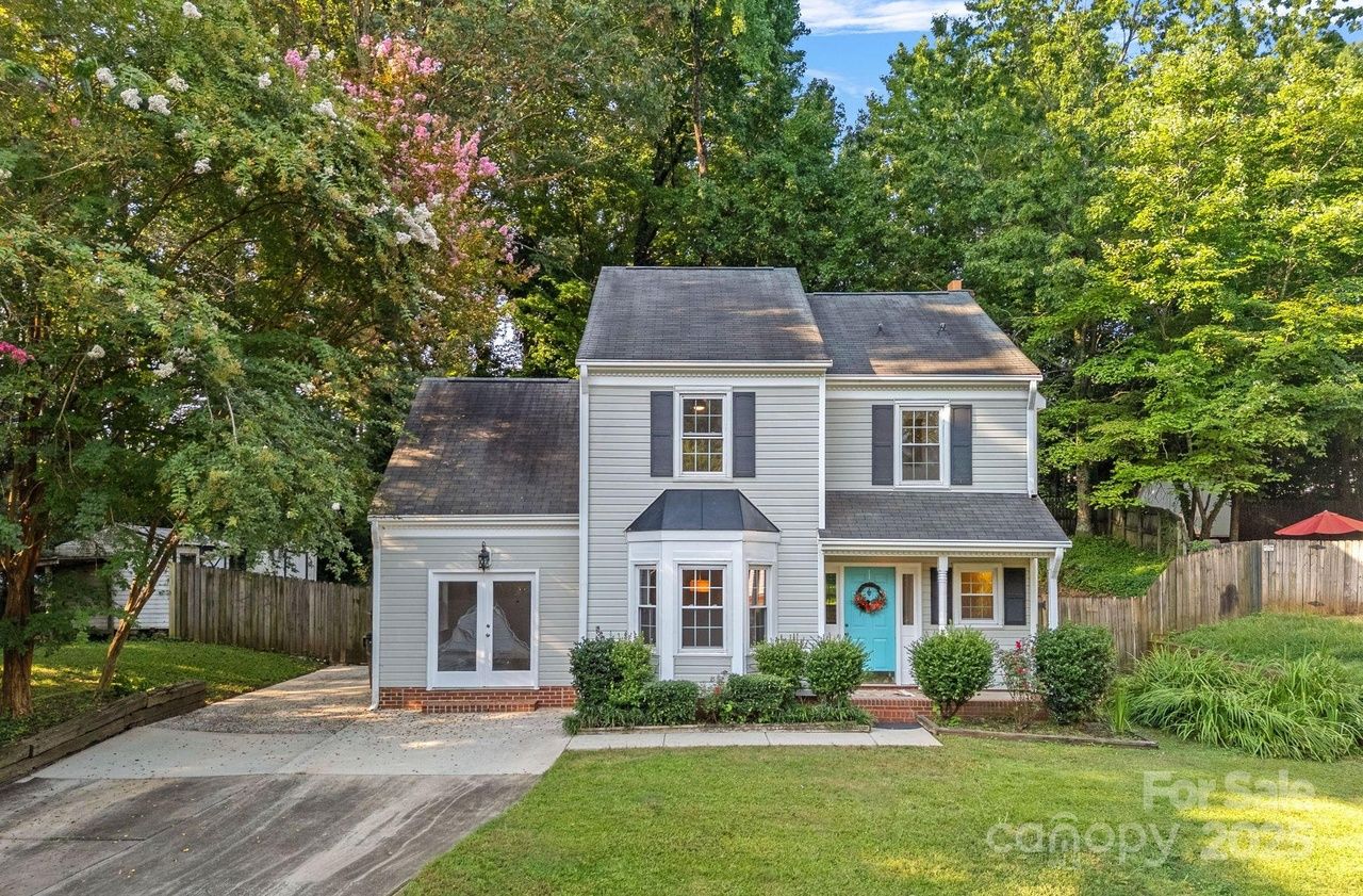 Two-story gray house with white trim, a bright blue front door with a wreath, and a bay window. Surrounded by green trees and bushes with a concrete driveway and lawn in front.