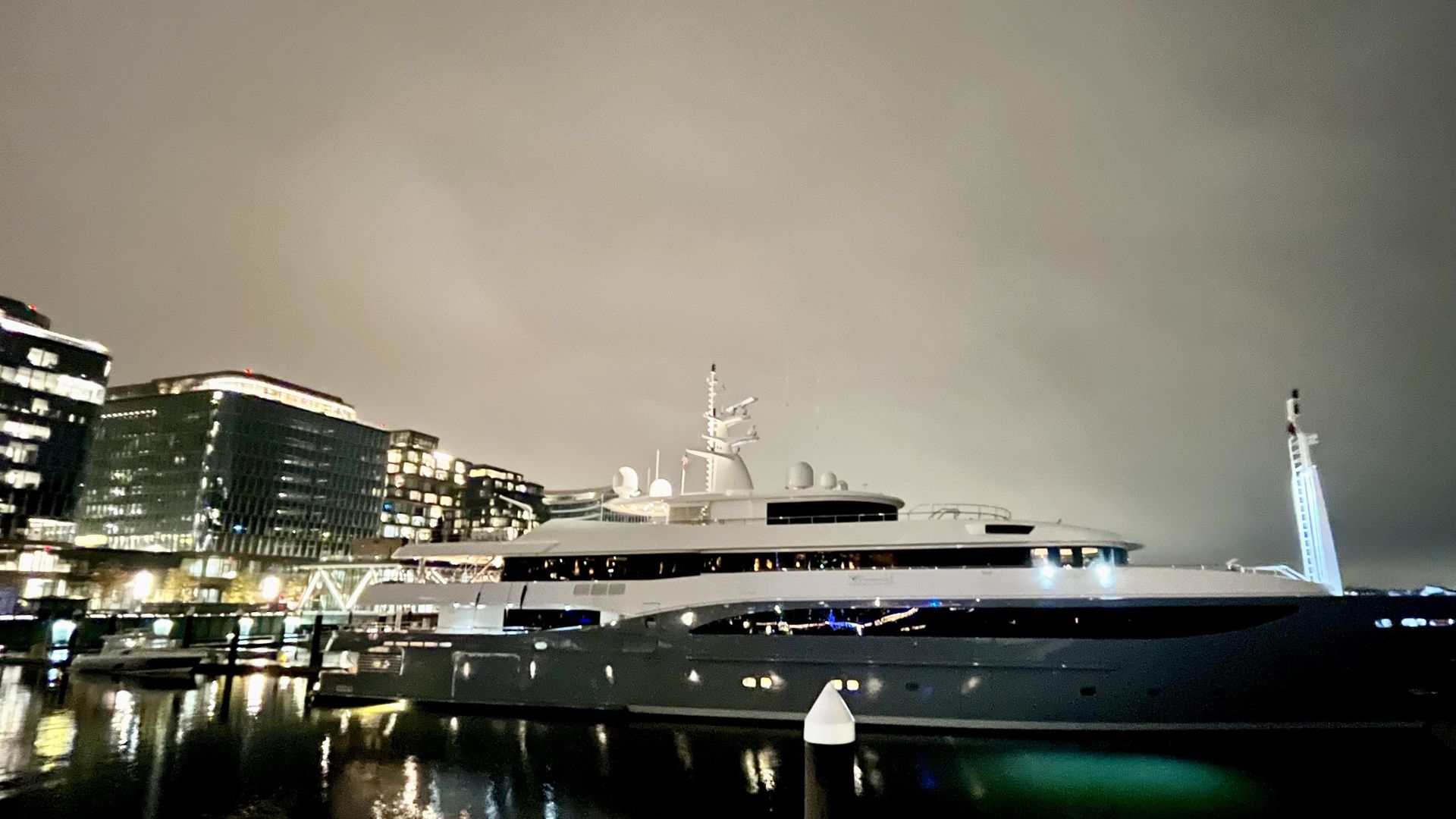 A picture of a superyacht docked at the Wharf at night, with lit buildings in the background