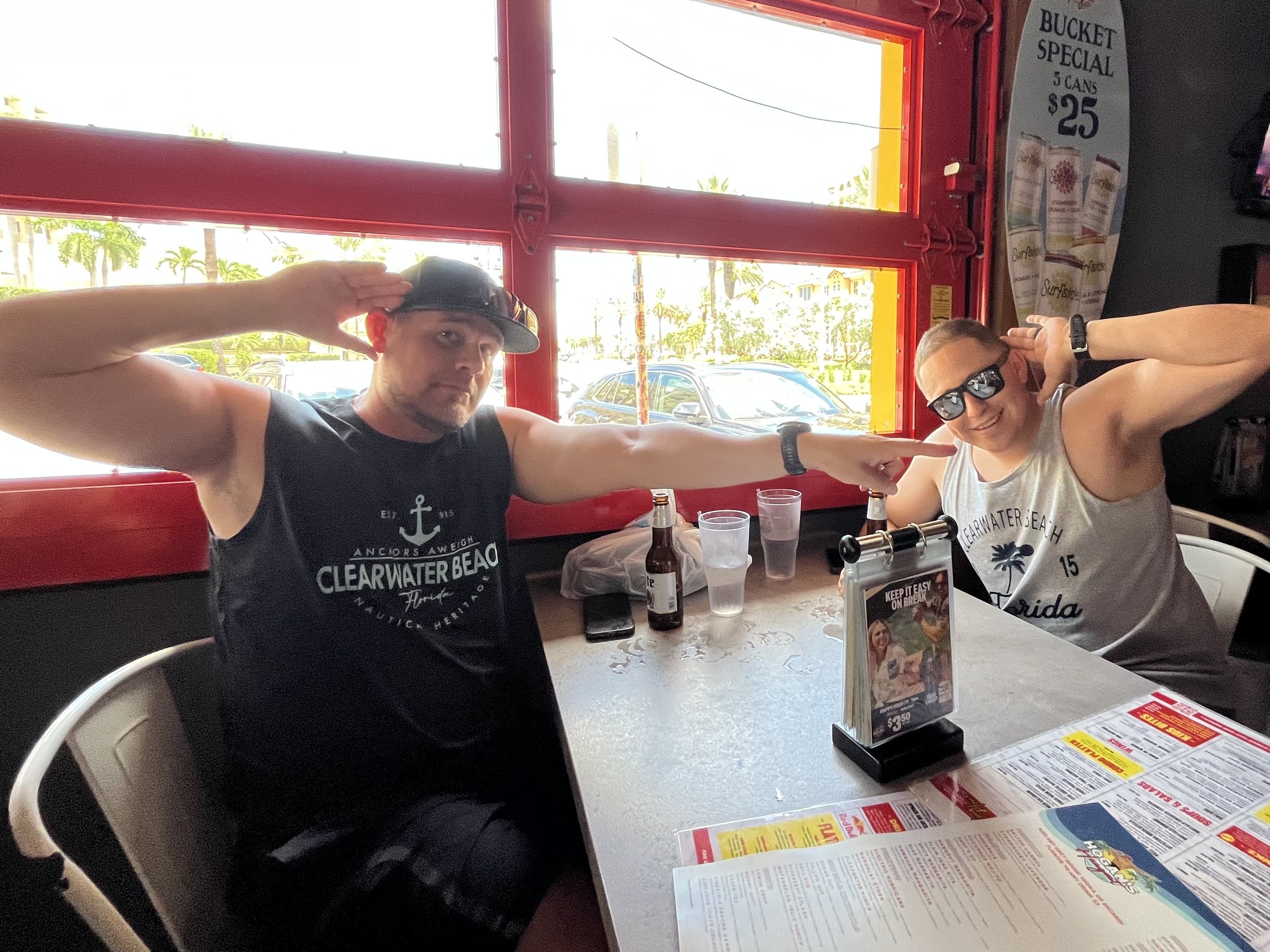 Two men sitting at a table inside a bar with red window frames, smiling and posing; one wearing a black sleeveless Clearwater Beach shirt and cap, the other in a gray tank top and sunglasses.
