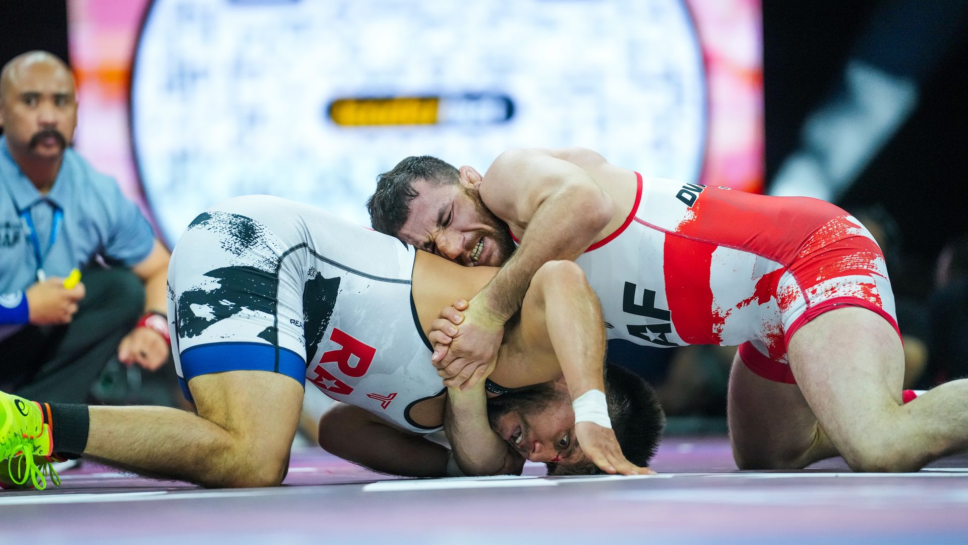 Two wrestlers in red and white singlets grapple on a mat; one pins the other as a blue-clad referee watches from the left, with a bright arena backdrop.