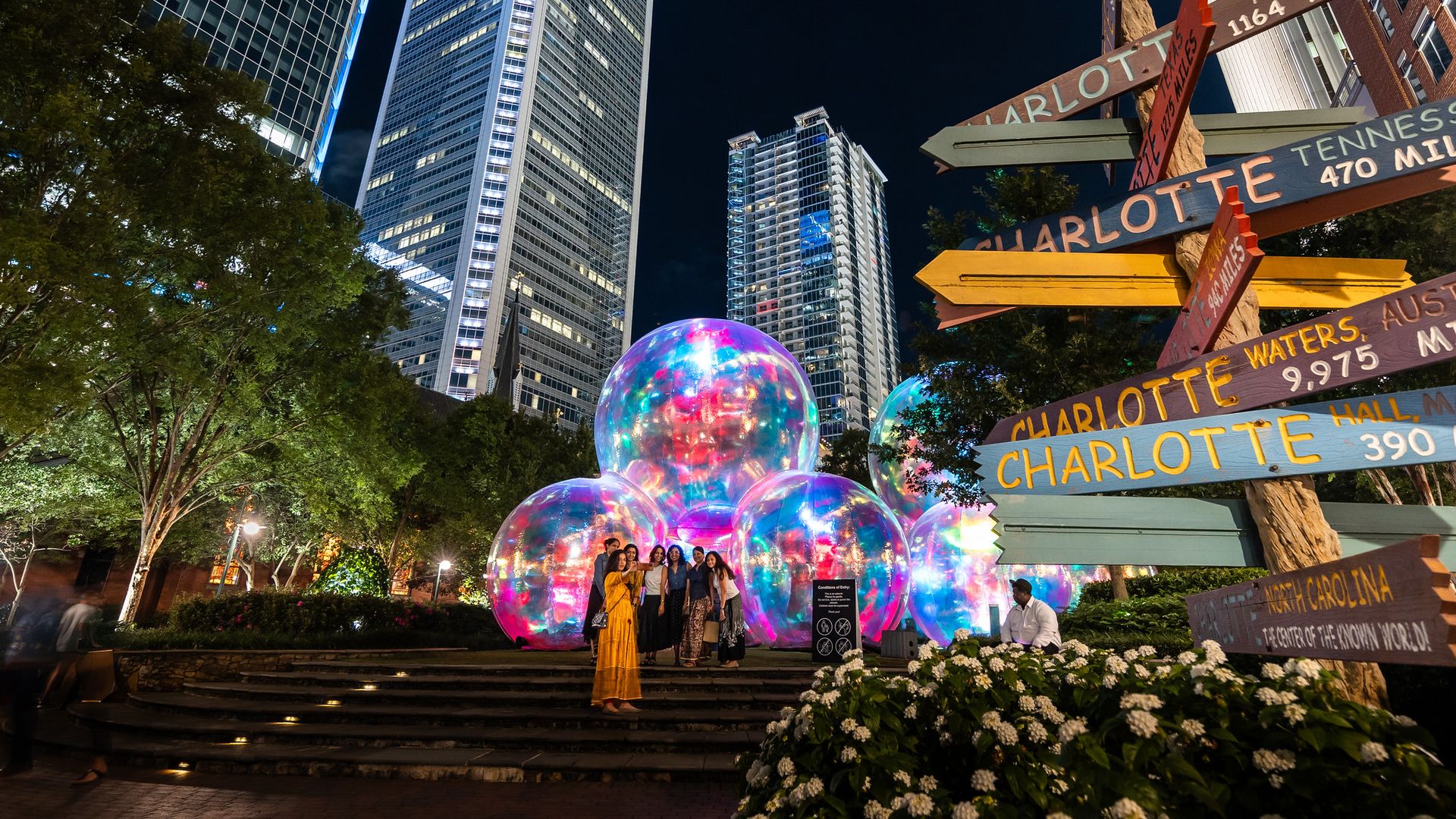 Night scene in a city park with tall illuminated skyscrapers, colorful glowing bubbles installation, a group of people posing, and a multi-directional wooden signpost with locations and distances.