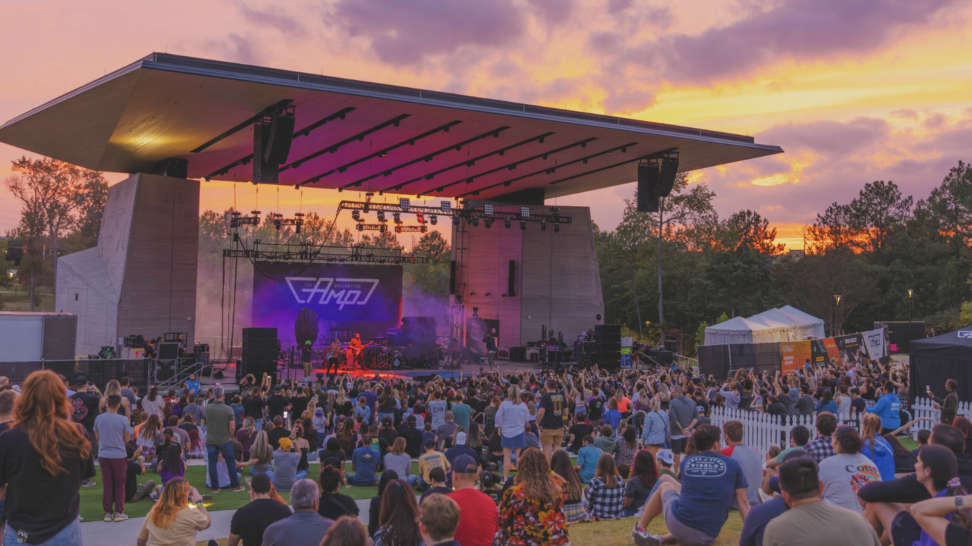 Outdoor concert at sunset with colorful sky, crowd watching band on stage under large roof, purple and orange lighting, trees and tents in background