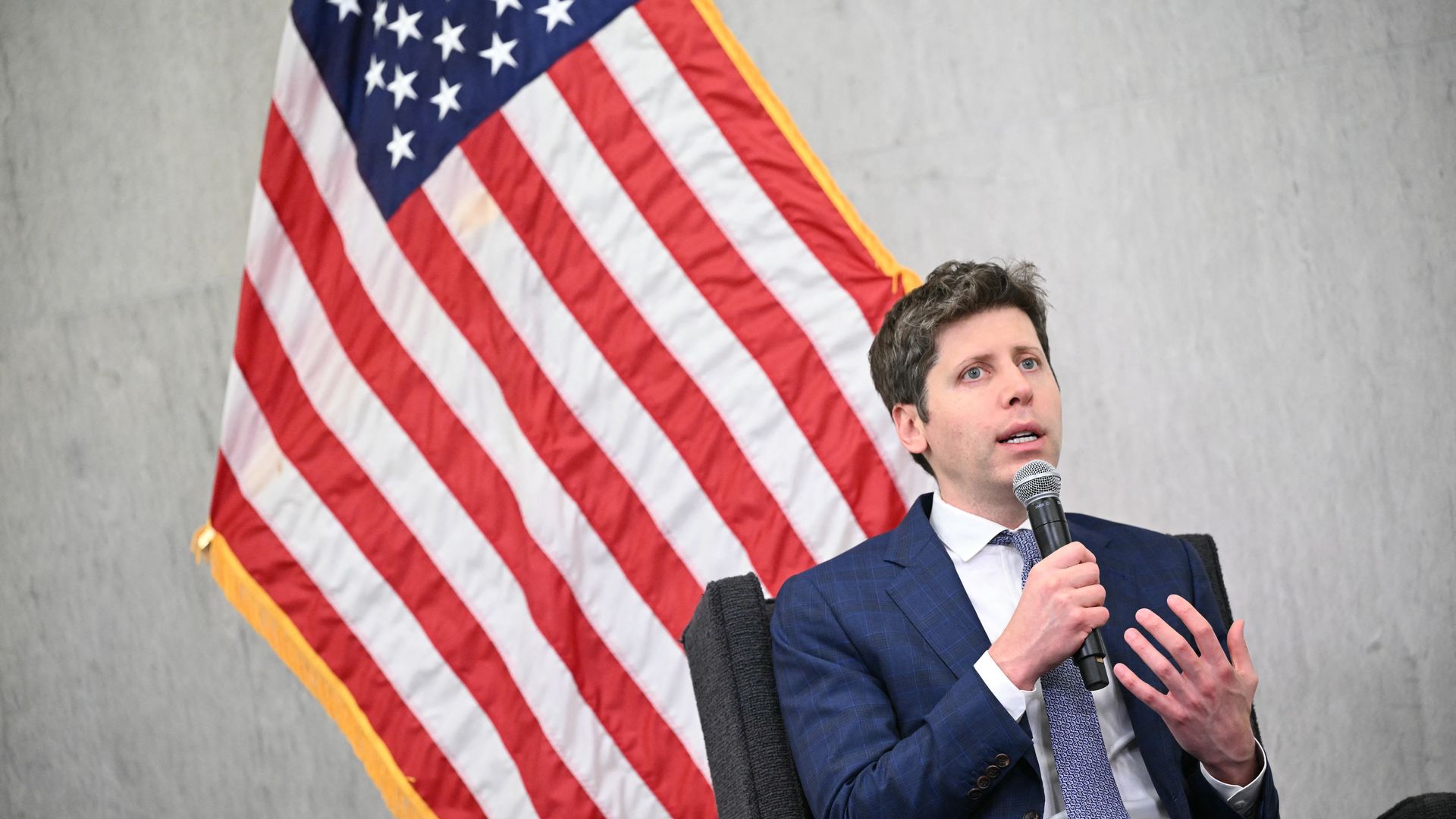 Man in a blue suit and patterned tie speaks into a microphone, seated in front of a large American flag with red, white, and blue stars and stripes on a grey background.
