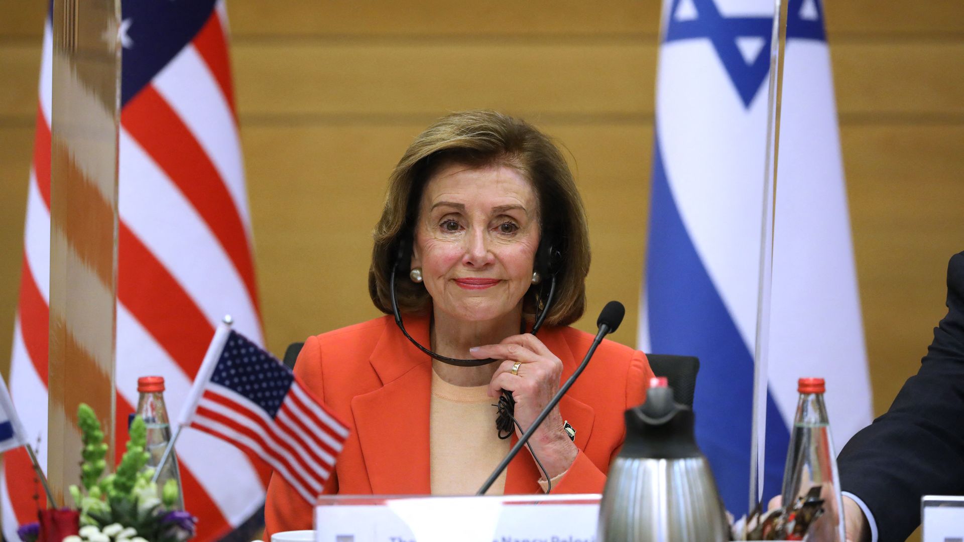 Former House Speaker Nancy Pelosi, wearing an organge blazer and tan blouse and taking off a headset, sits in front of a microphone flanked by U.S. and Israeli flags.