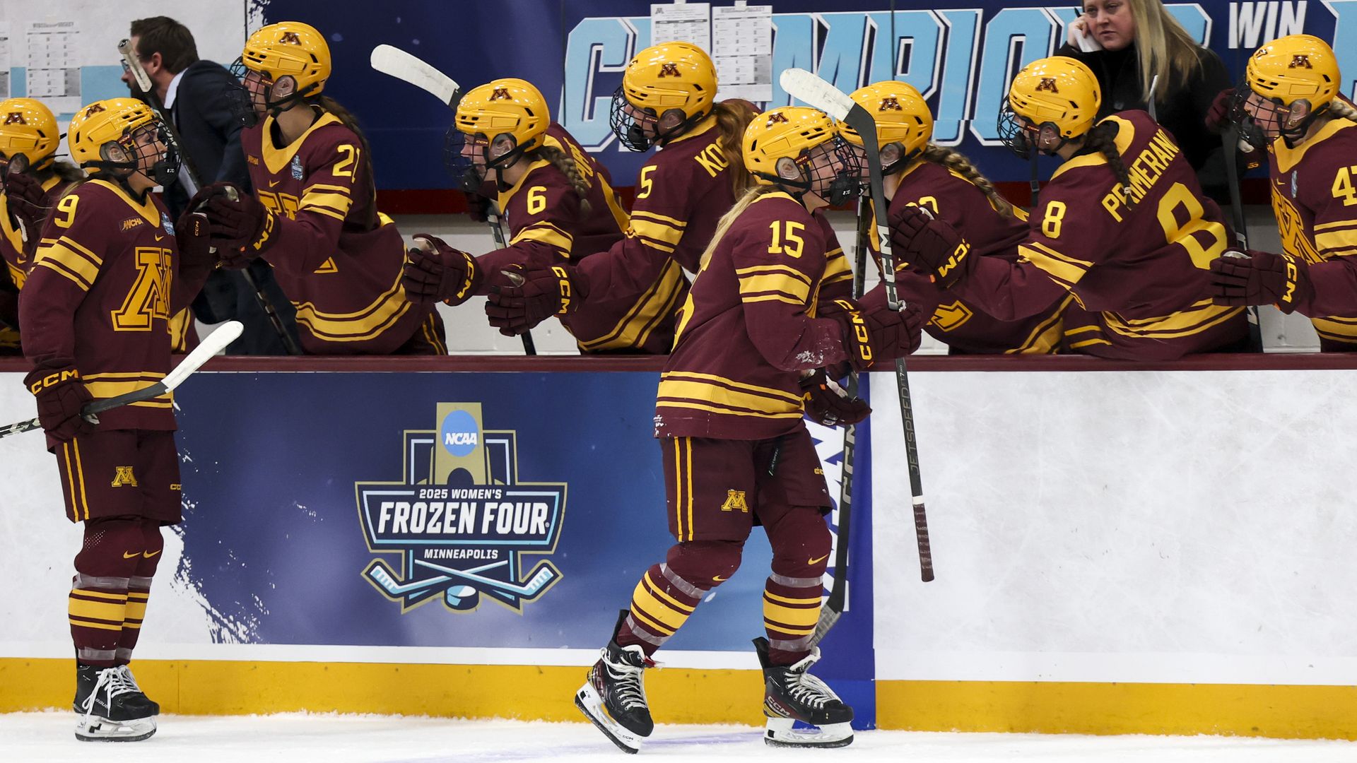 Minnesota women's hockey players in maroon and gold uniforms with yellow helmets high-five along the boards, sticks raised; blue "Frozen Four" banner and cheering crowd in background.