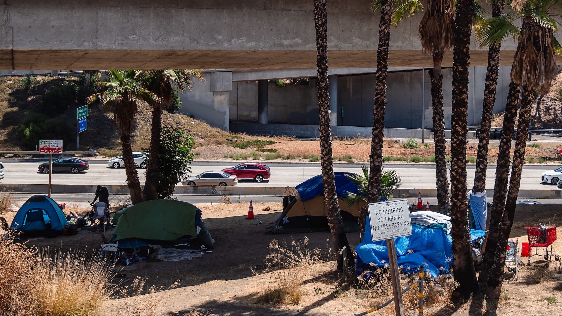 Four tents and a shopping cart sit near an overpass at a homeless encampment.