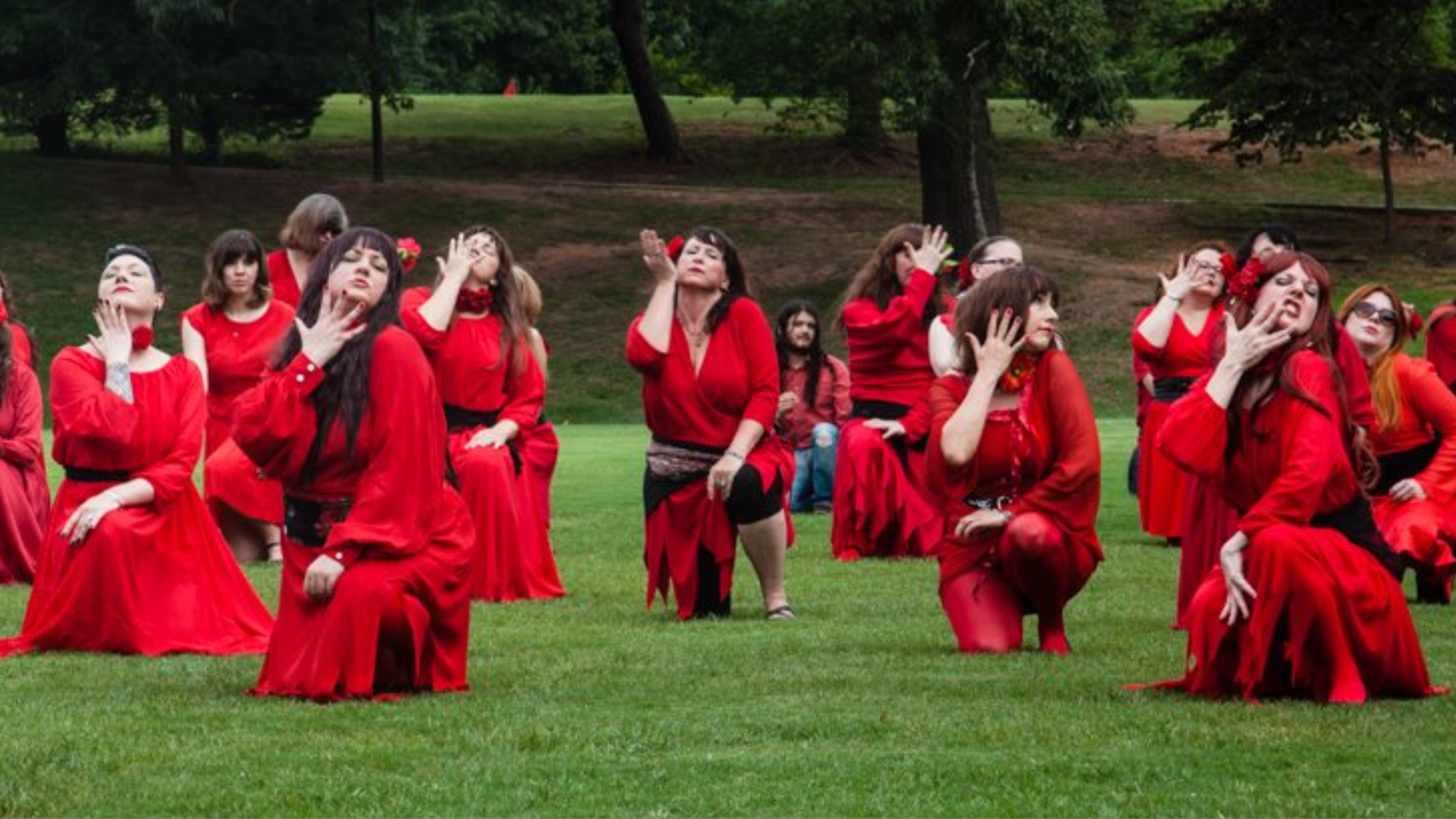A group of women in a public park wearing red dresses crouch on one knee and put their hands to their faces to recreate a Kate Bush music video