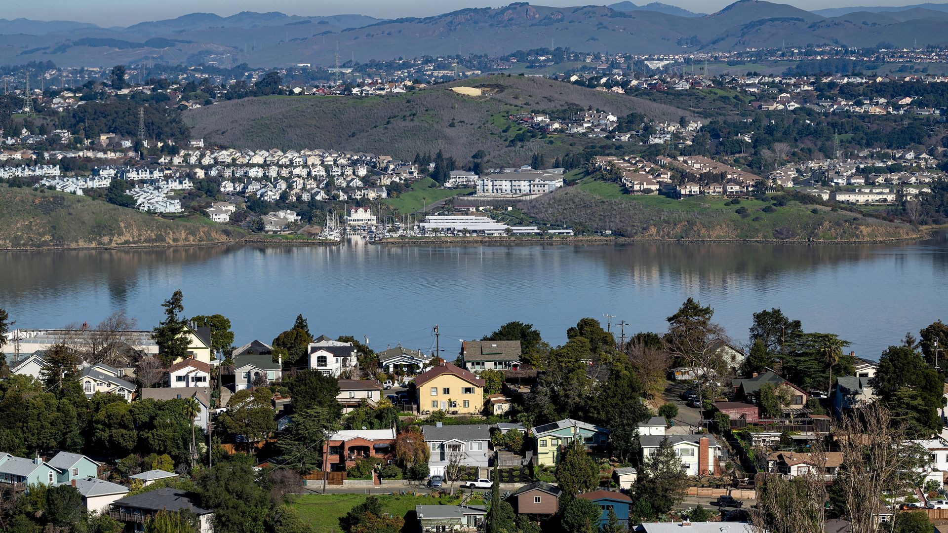 Suburban neighborhood with various houses and trees in foreground, calm body of water in middle, and hills with more houses under blue sky in background.