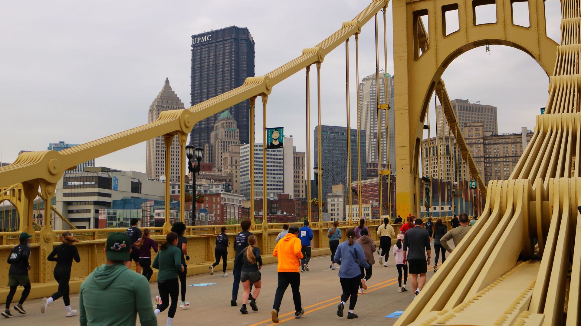 Runners jog across a bright yellow suspension bridge in Pittsburgh; a downtown skyline features a tall blue UPMC building visible in the background.