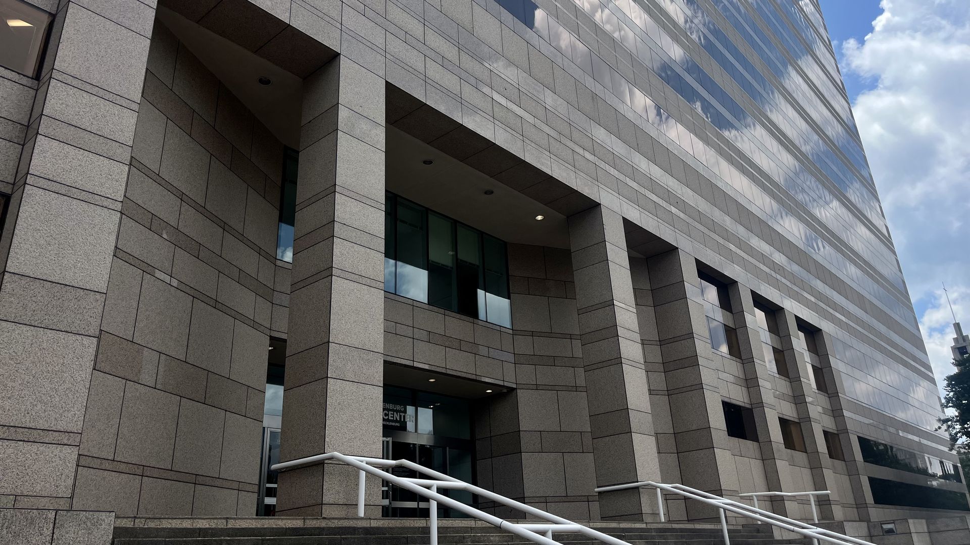 Tall modern building with a gray stone facade and glass windows, wide staircase with white railings leading up to the entrance under an overhang on a partly cloudy day.