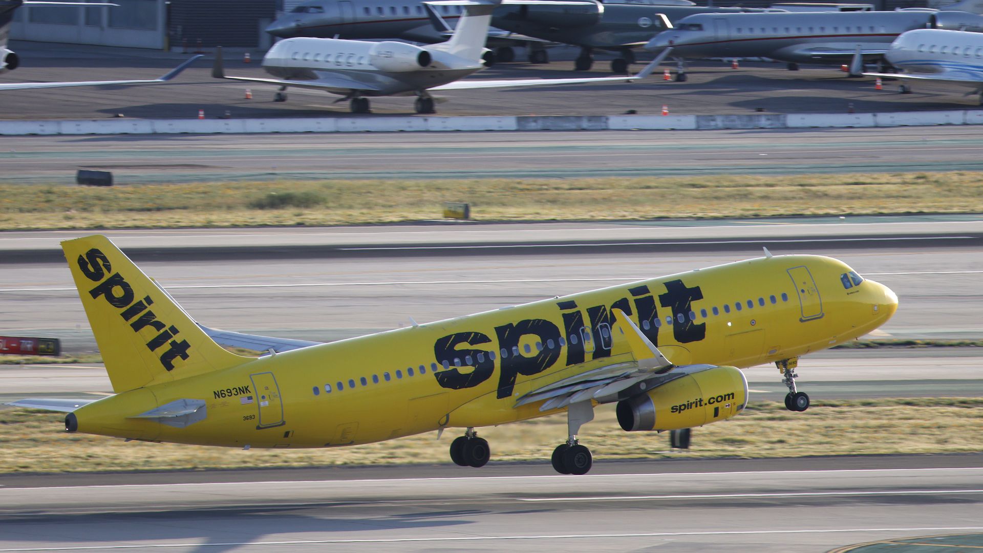 Yellow Spirit Airlines airplane taking off on runway, with black "Spirit" text on fuselage and tail, airport tarmac and several private jets in background.