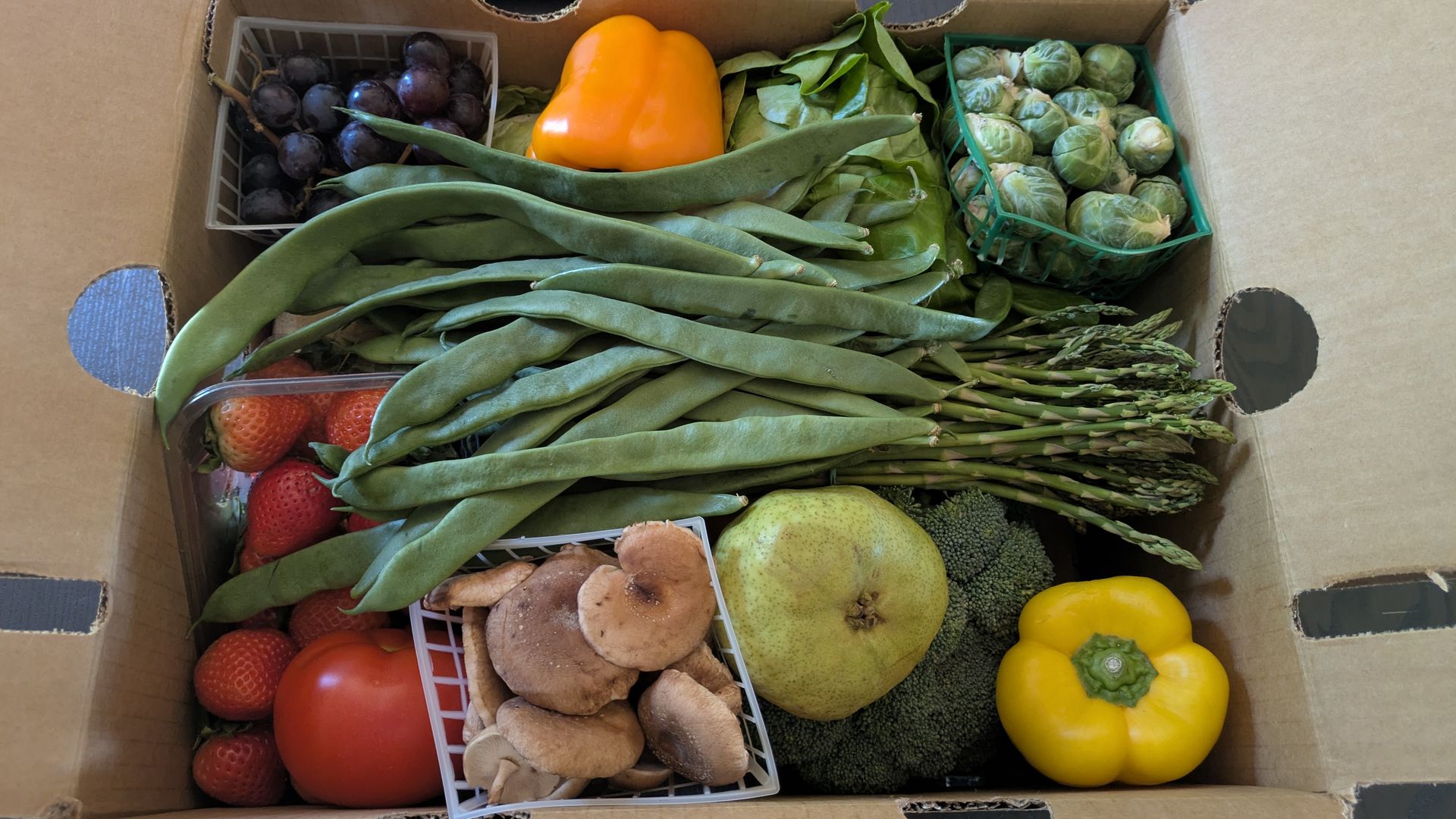 Top-down view of an open cardboard box with assorted produce: orange bell pepper, long green beans, Brussels sprouts, green grapes, yellow pepper, tomatoes, strawberries, mushrooms, and a large green fruit.