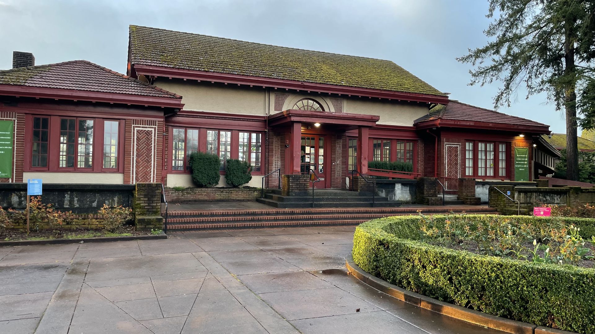 Historic brick and stucco building with red trim and moss-covered roof, surrounded by trimmed hedges and rose bushes. A ramp and stairs lead to the main entrance under a small portico.