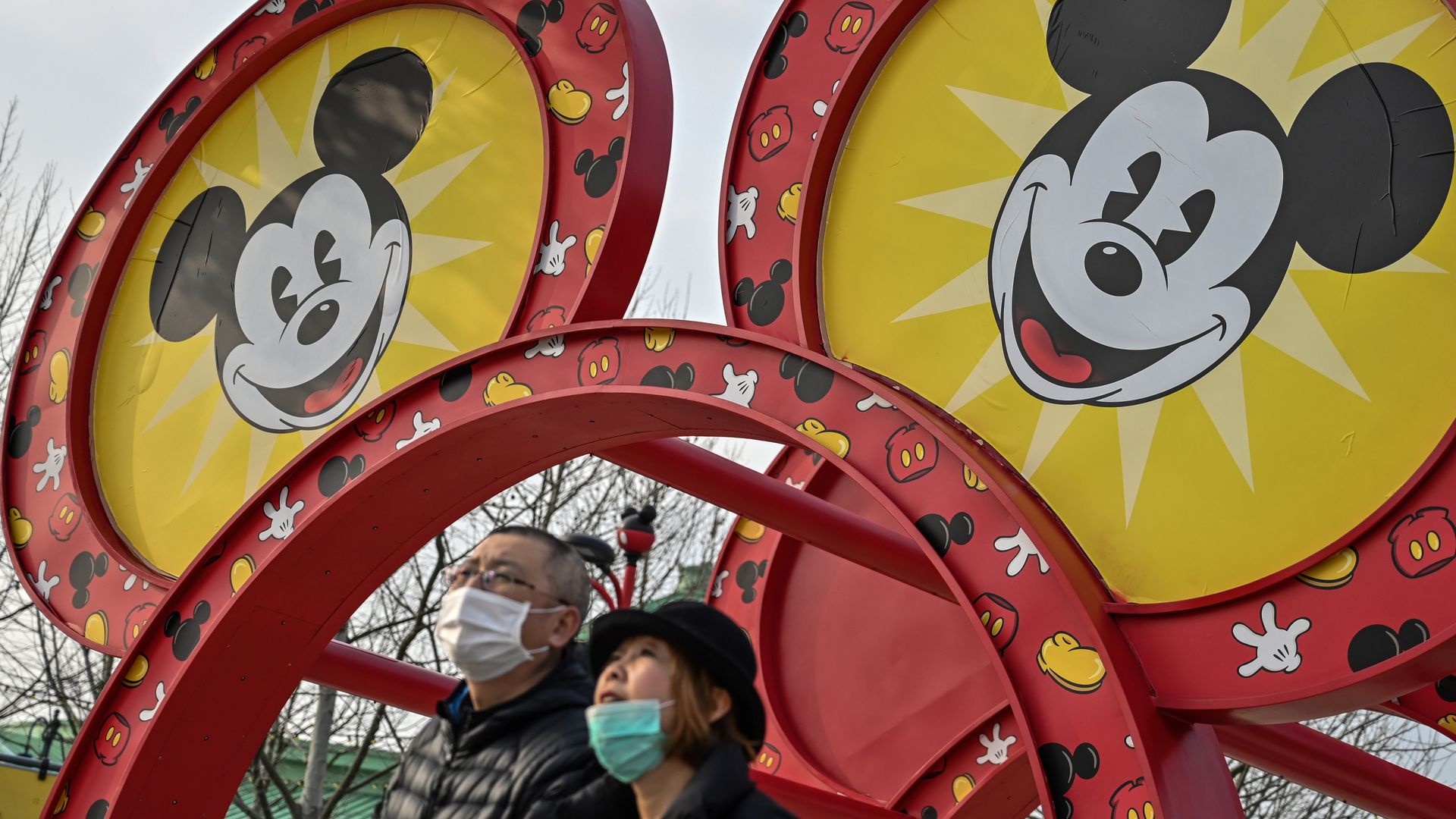 In this image, two people wearing face masks stand underneath a giant Mickey Mouse