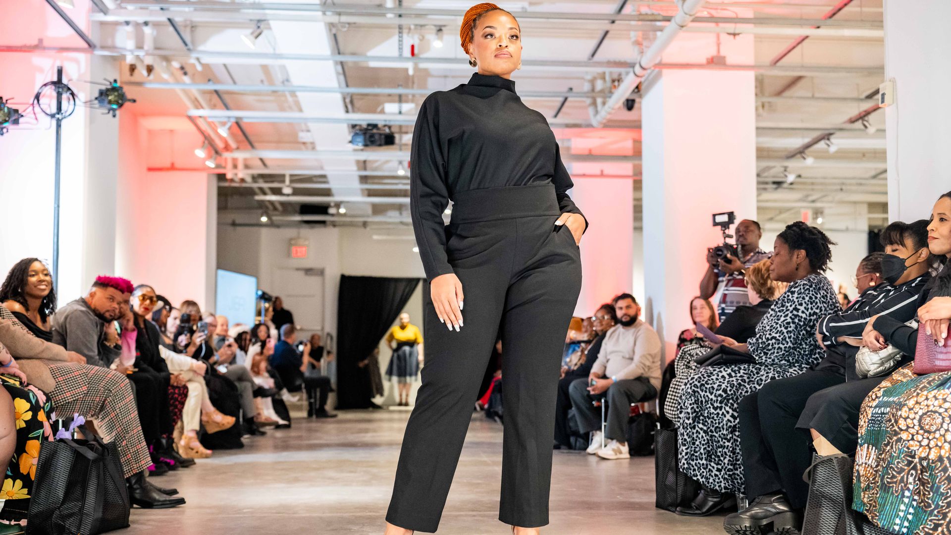 Confident model with braided orange hair in black outfit and black heels walking on runway at fashion show, with a seated audience watching on both sides.
