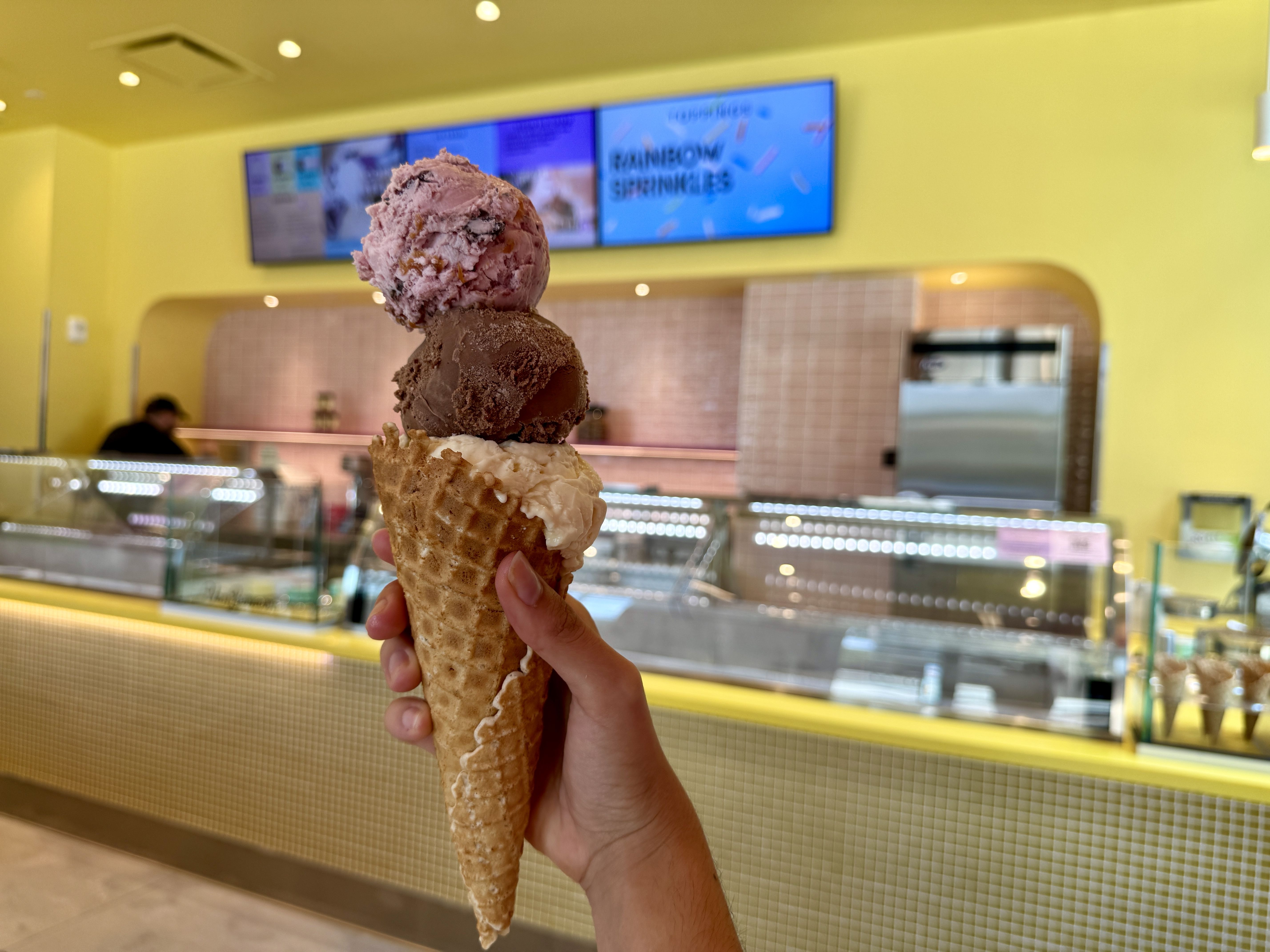 Hand holding a waffle cone with three scoops of ice cream in pink, brown, and off-white colors inside a yellow-themed ice cream shop.