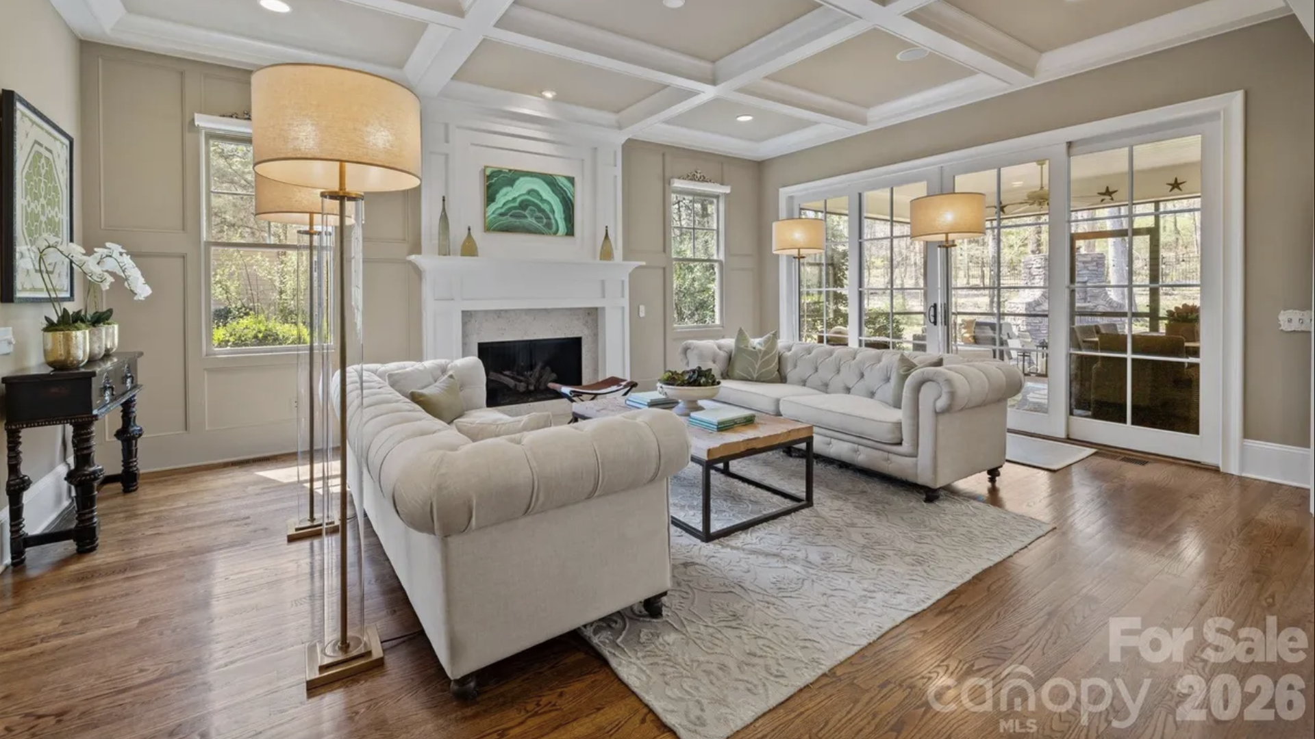 Bright living room with a coffered ceiling, beige walls, white fireplace mantel, and two tufted white sofas around a wooden coffee table. Hardwood floors and a light rug with glass doors to a patio.