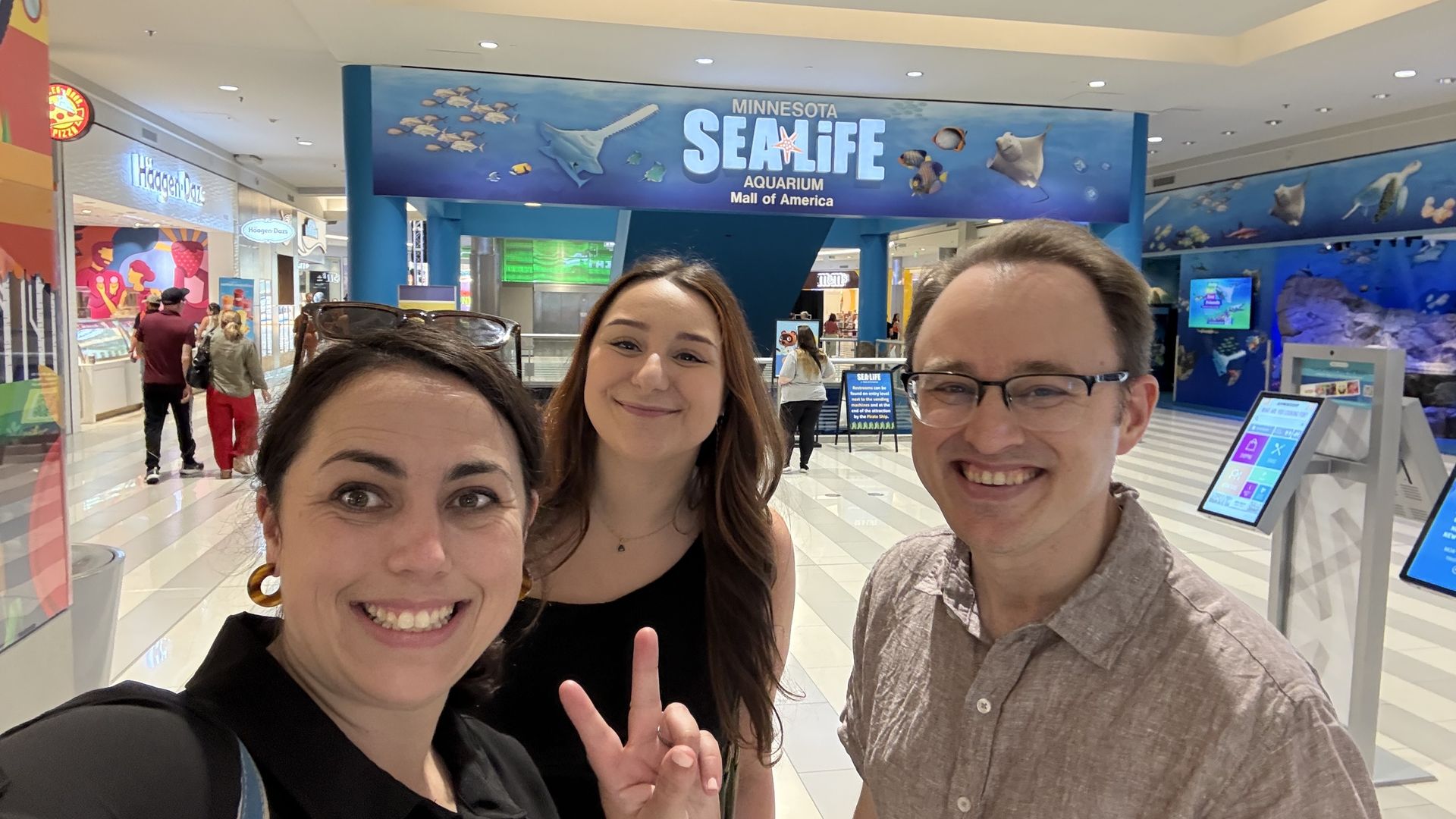 Three friends take a cheerful selfie in a mall by a large blue banner reading "MINNESOTA SEA LIFE" and "Mall of America"; Haagen-Dazs shop visible in the background.