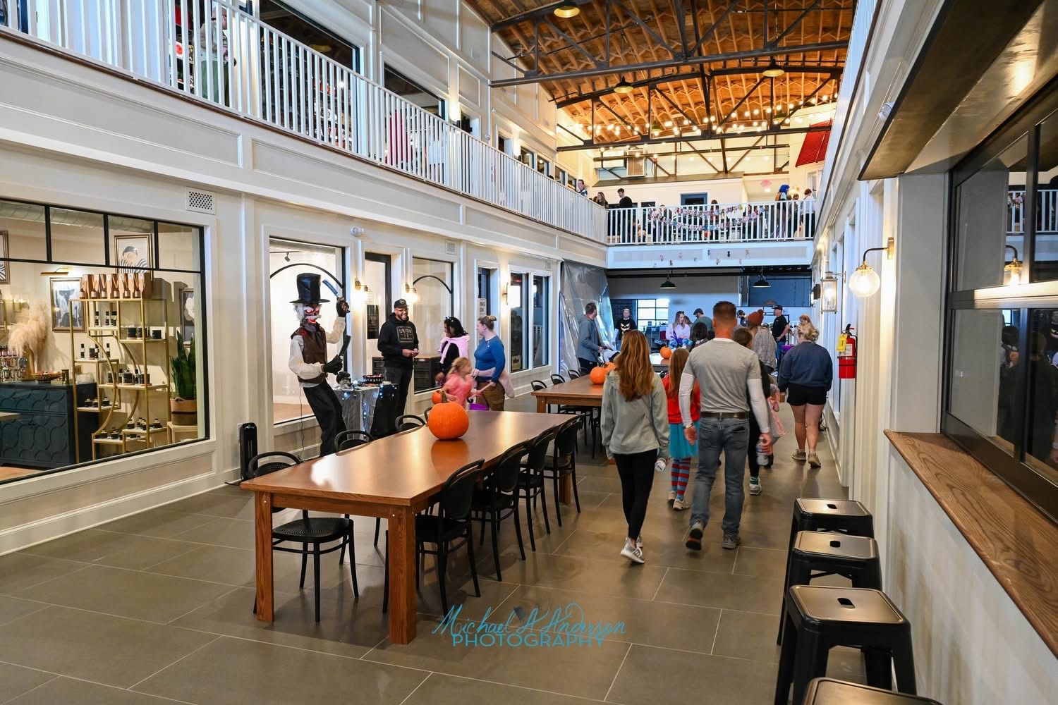 Indoor market or event space with wooden tables, black chairs, pumpkins on tables, people walking, a performer in a tall hat and mask, and two levels with white railings under a wooden ceiling.