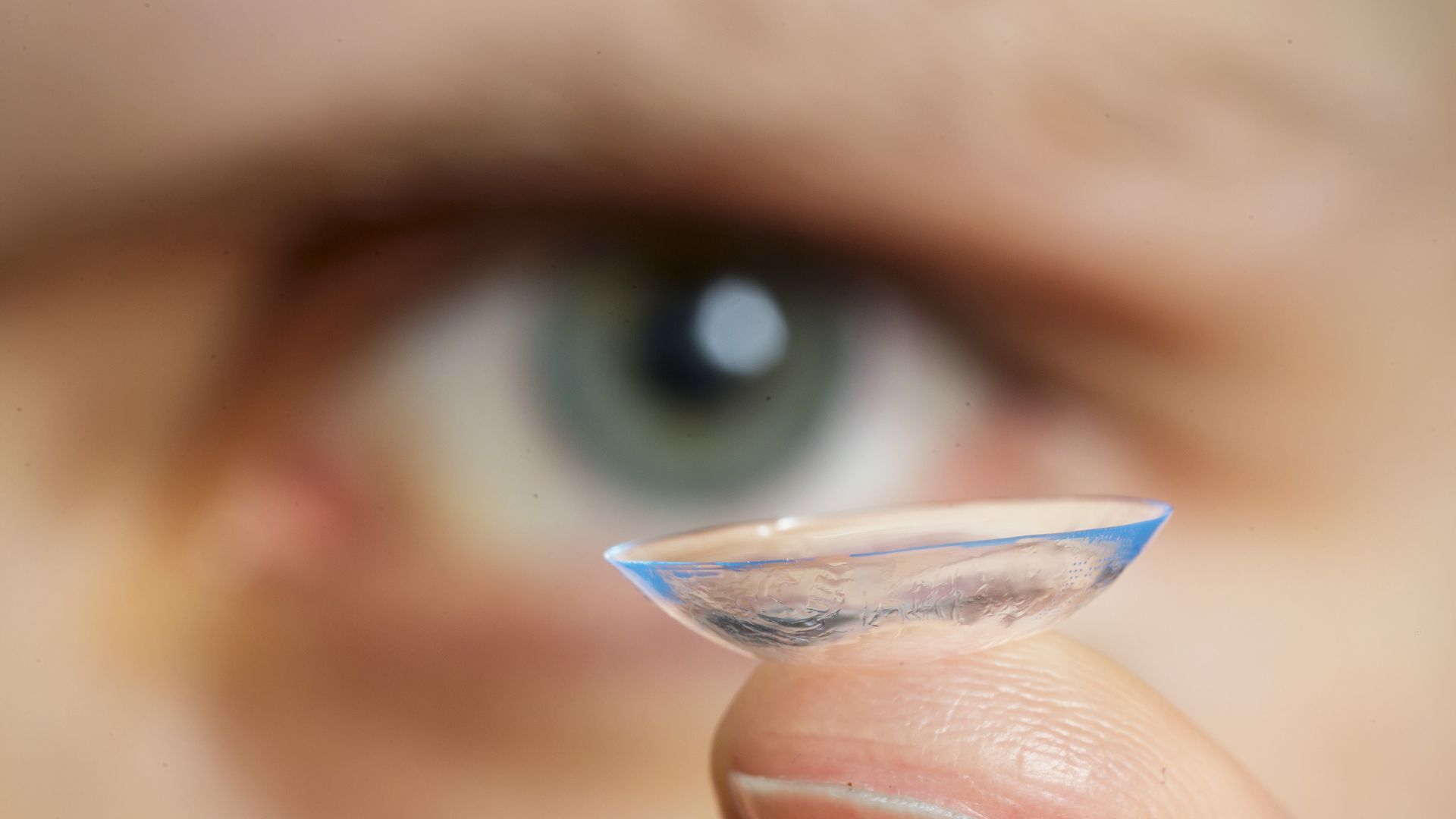 Someone holding up a contact lens on their finger with a blurred blue eye in the background