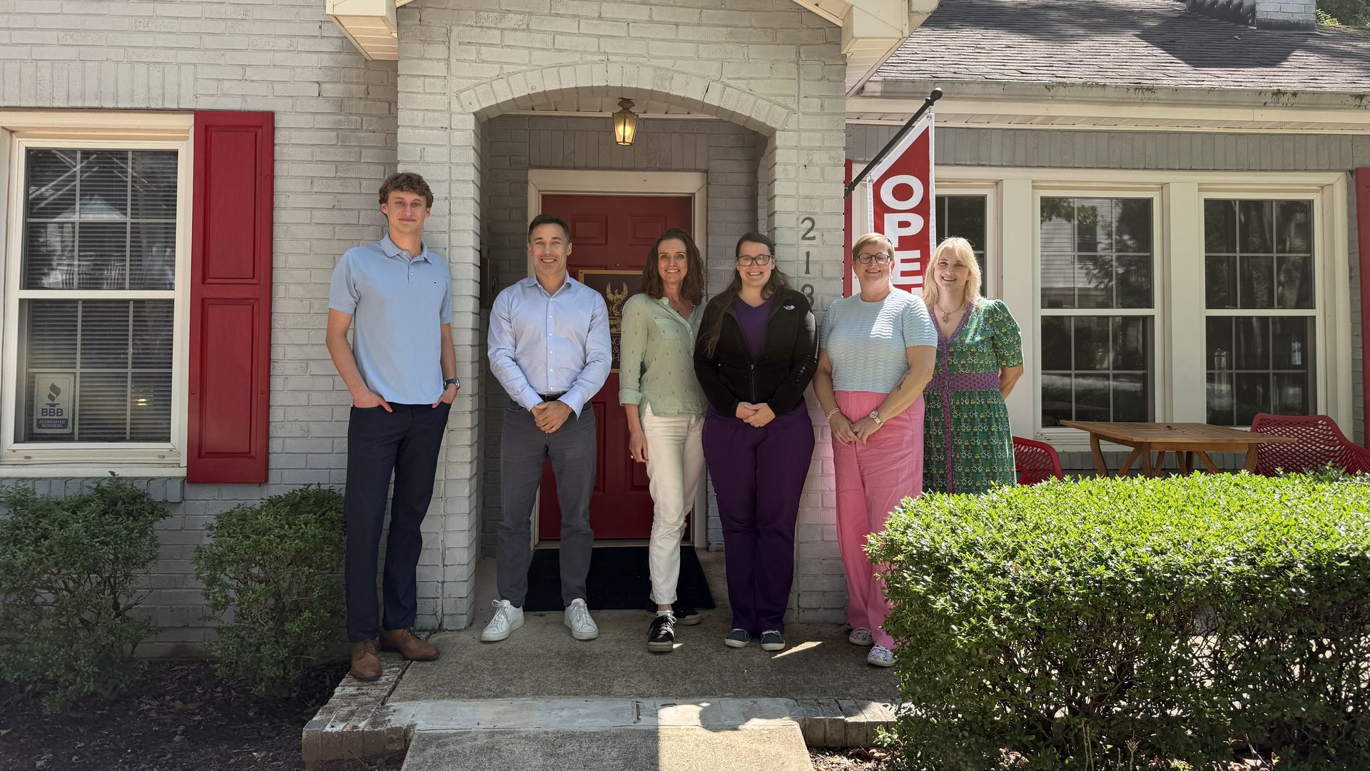 Several people stand on the front porch of a light gray brick house with a red door under a gabled entry. A red and white "OPEN" flag, greenery, and flowering bushes are visible.