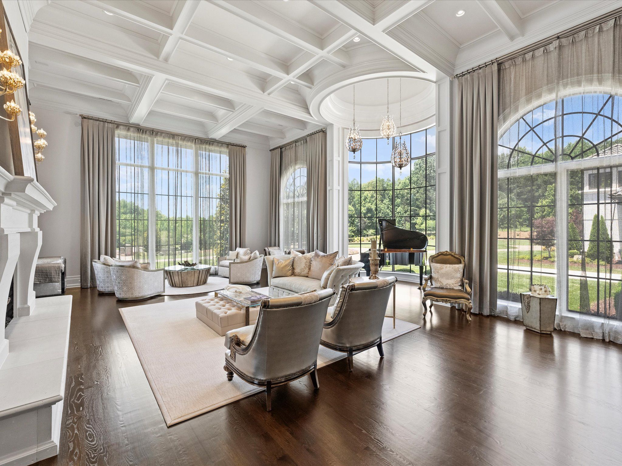 Bright, spacious living room with white coffered ceiling, large arched windows with sheer curtains, light-colored furniture, grand piano, and dark hardwood floors.