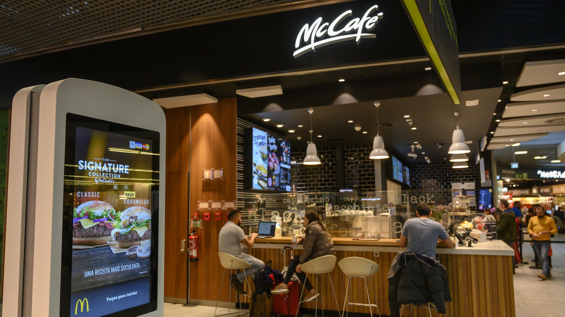 In this image, a man sits at a counter at a McCafe. In the foreground, a touch-screen stands upright for customers to order electronically. 