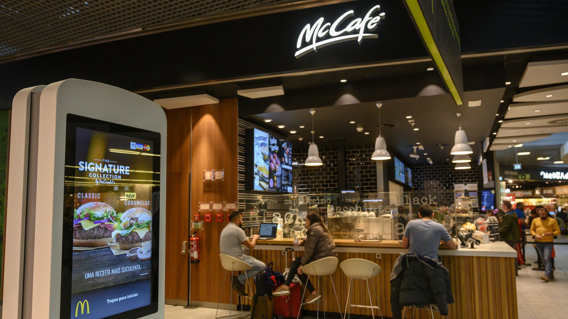 In this image, a man sits at a counter at a McCafe. In the foreground, a touch-screen stands upright for customers to order electronically.