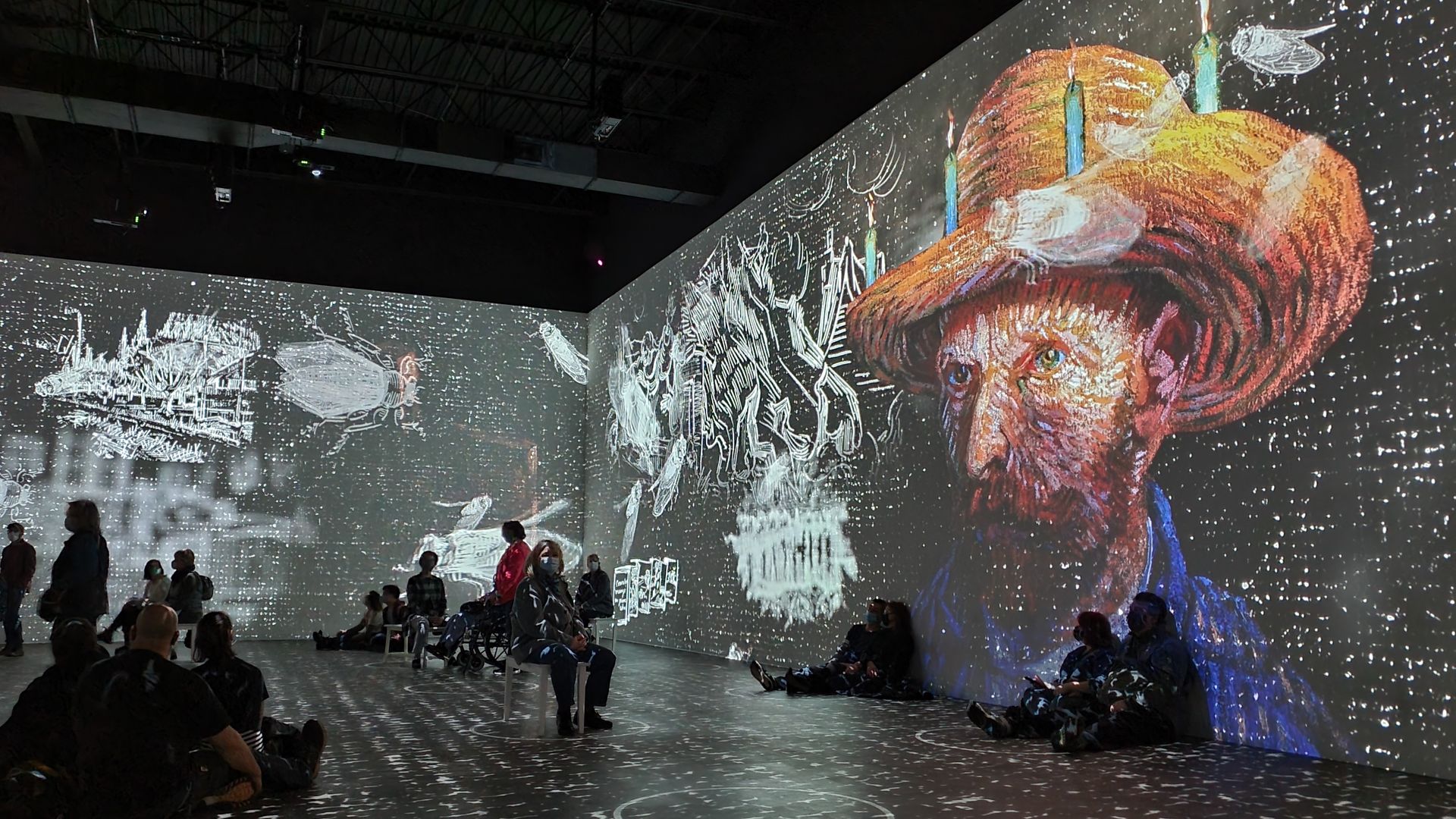 An immersive Van Gogh exhibit casts a photo of Van Gogh on a wall while people sit on benches or the floor observing it