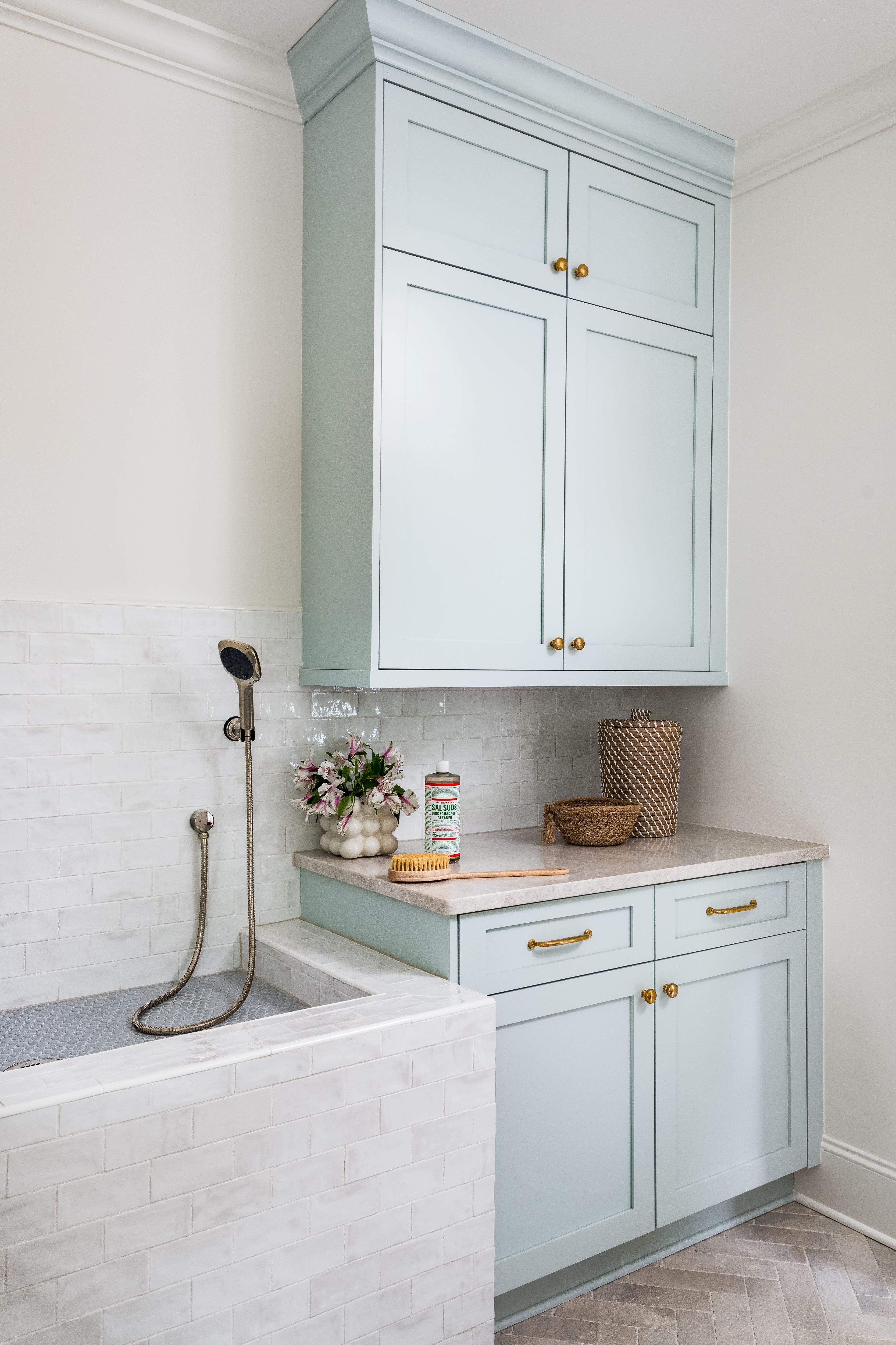 Light blue bathroom cabinetry with gold handles next to a white tiled shower with a handheld showerhead, a cleaning bottle, a plant in a round vase, and woven baskets.