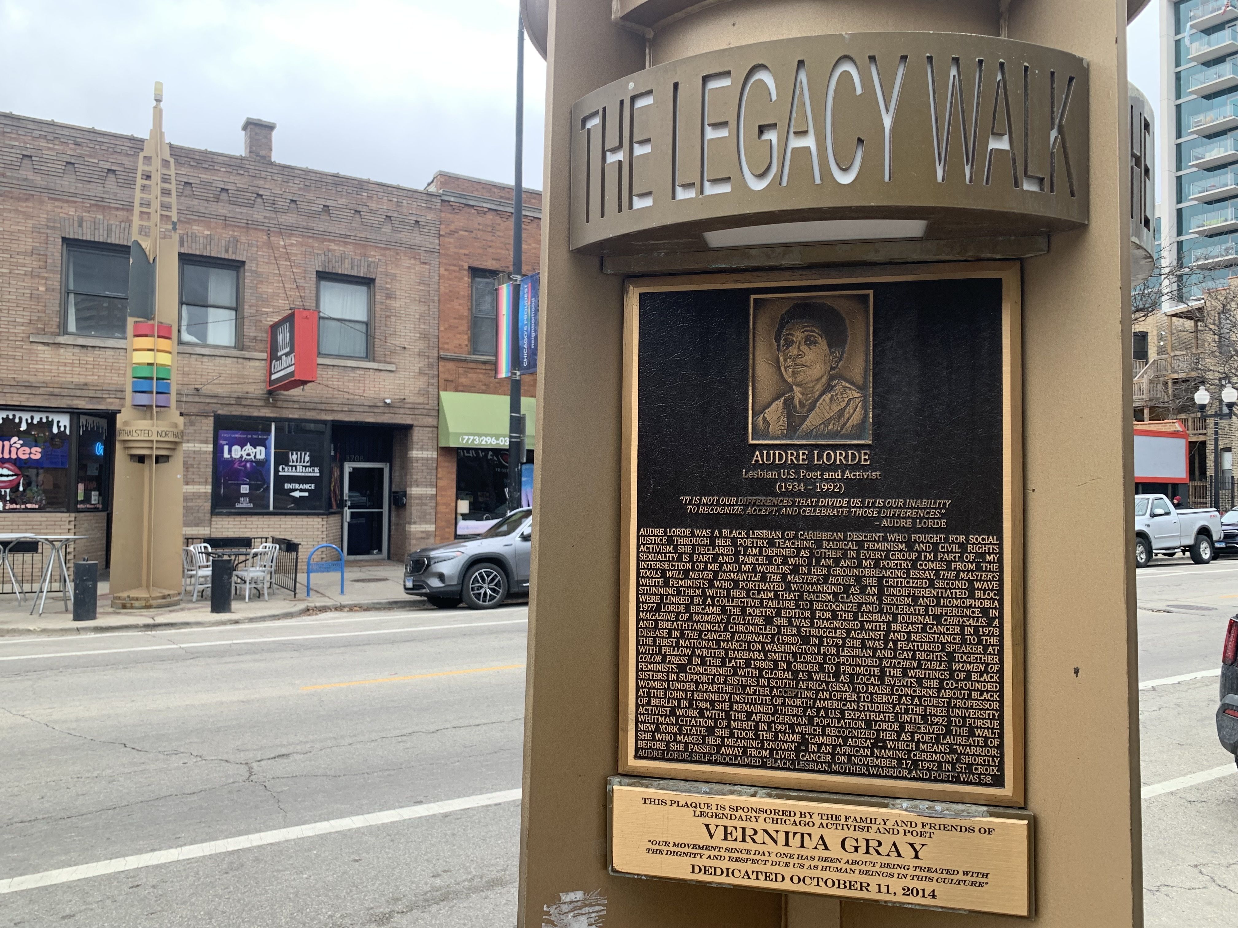 Bronze plaque on The Legacy Walk honoring lesbian U.S. poet and activist Audre Lorde, with detailed biography, mounted on a brown pillar on a city street with rainbow flags and buildings in the background.