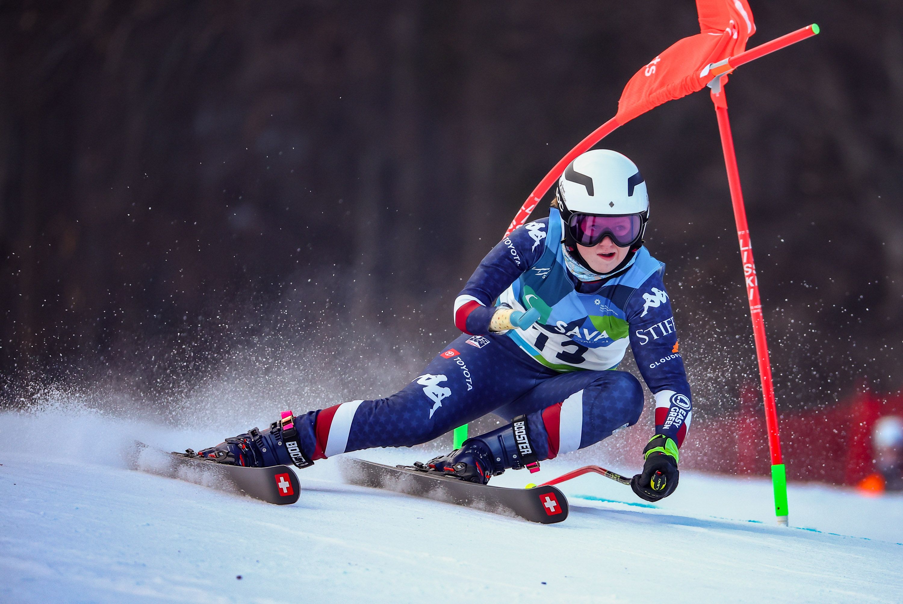 Skier in blue race suit with number 13 and white helmet speeds down snowy slope, leaning sharply near red gate flag with snow spraying behind skis marked with white cross.