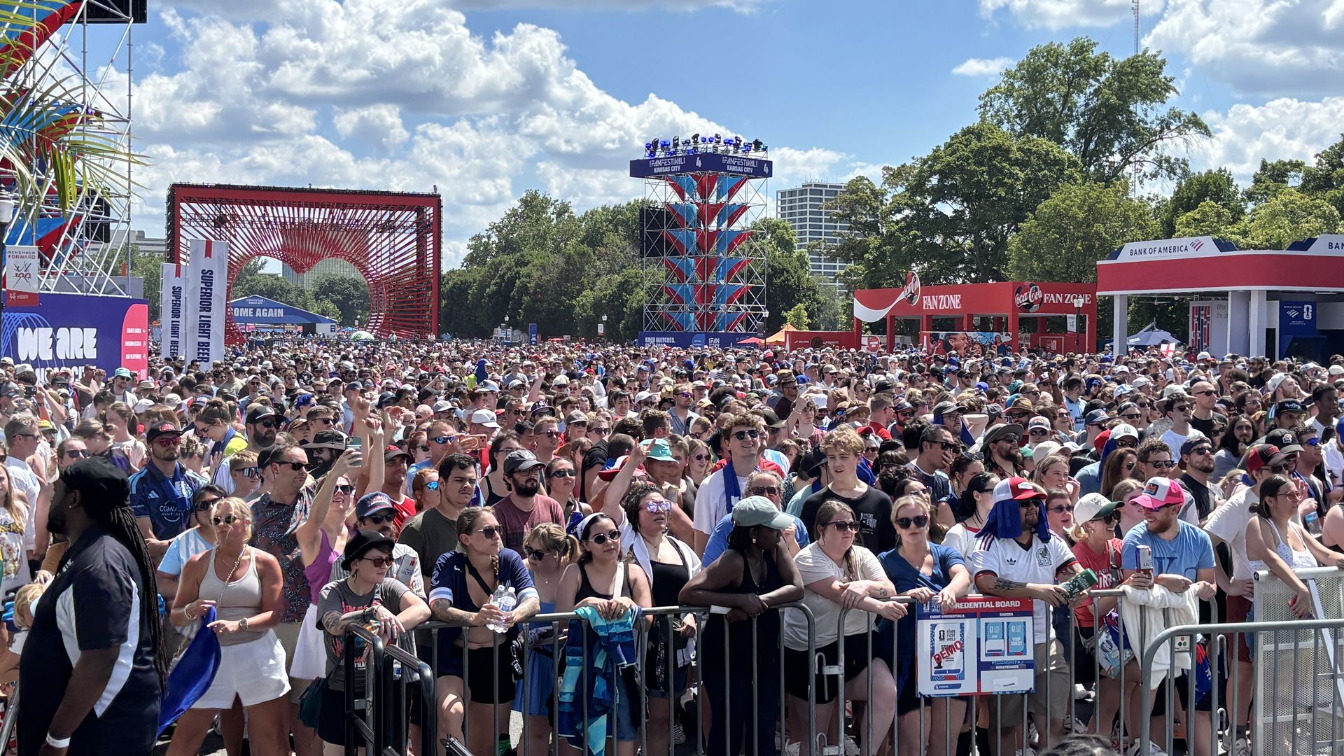 A large outdoor festival crowd stands behind barriers, many wearing sunglasses and hats. In the background are colorful stage towers, a red sculptural arch, banners, trees, and a bright blue sky.