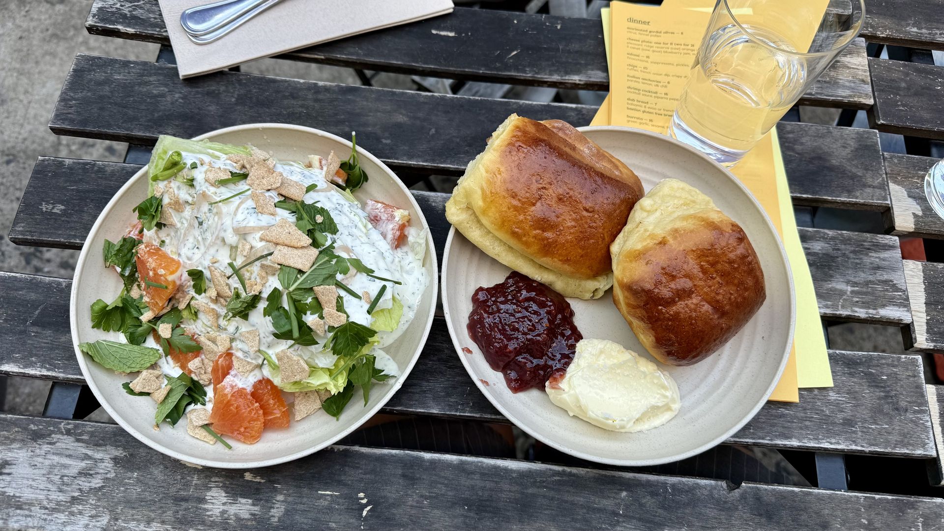 Two plates on a rustic wooden table: one with a fresh salad topped with herbs and crispy bits, the other with two golden brown rolls, a dollop of cream, and a spoonful of red jam.