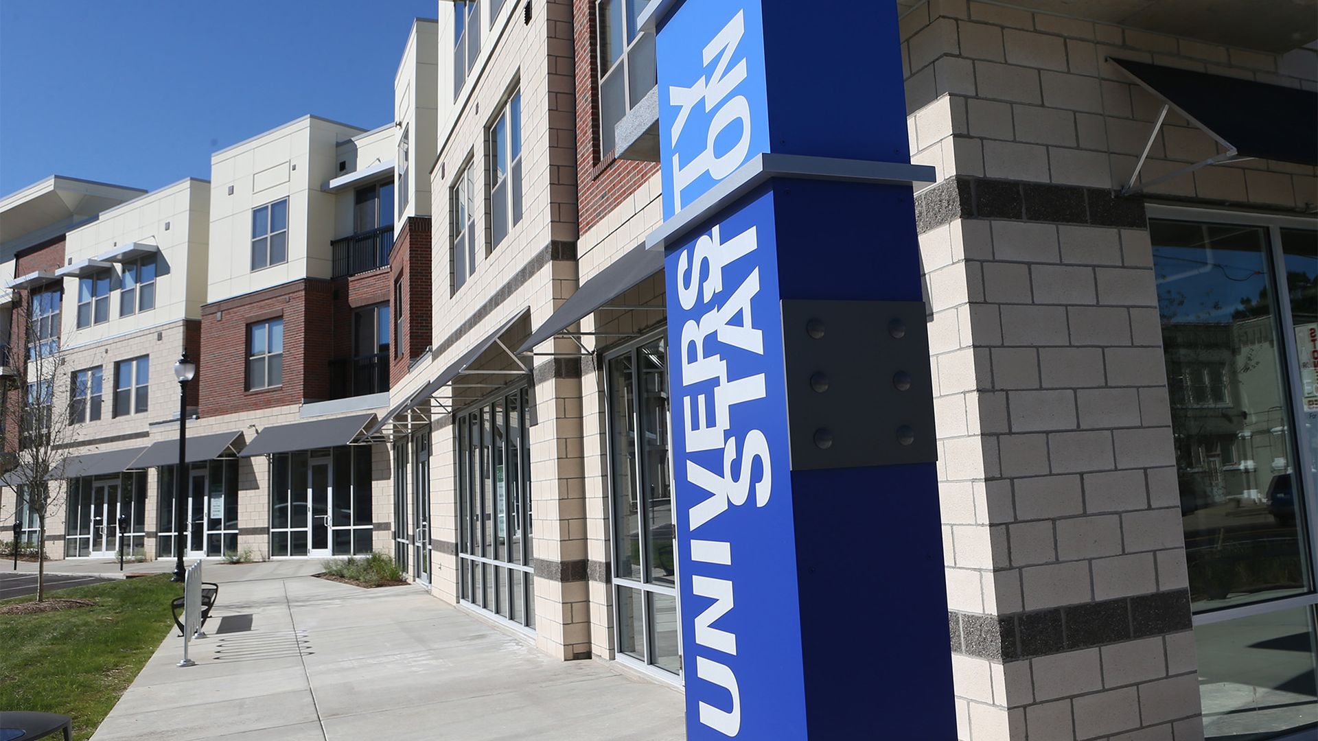 Modern storefronts with large glass windows and beige brickwork line a sunny sidewalk; a tall blue sign with white vertical letters stands at the corner, under a clear blue sky.