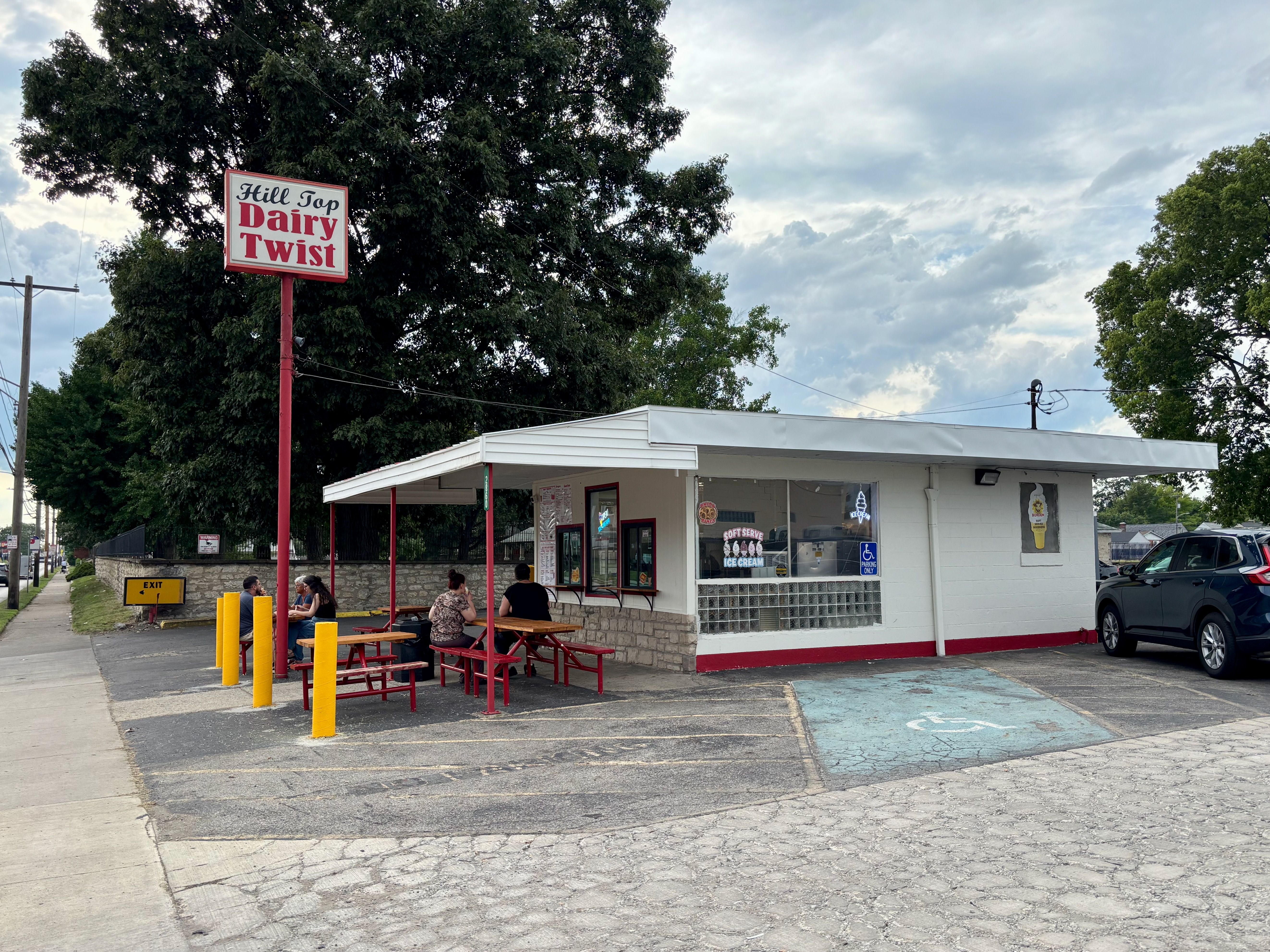 Outdoor diner with a tall red sign reading "Hill Top Dairy Twist" beside large trees. White building with big windows and red benches; several customers sit at picnic tables under a cloudy sky.