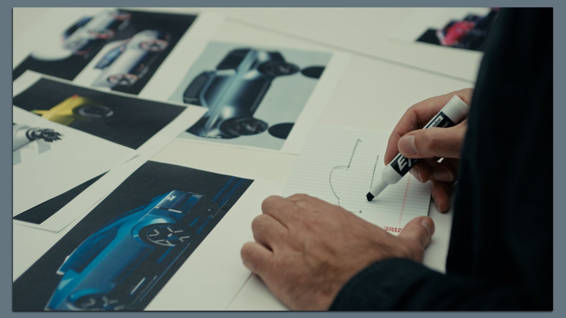 A person sketching a car design on a notepad with a black marker, surrounded by printed photos of various car models on a white table.
