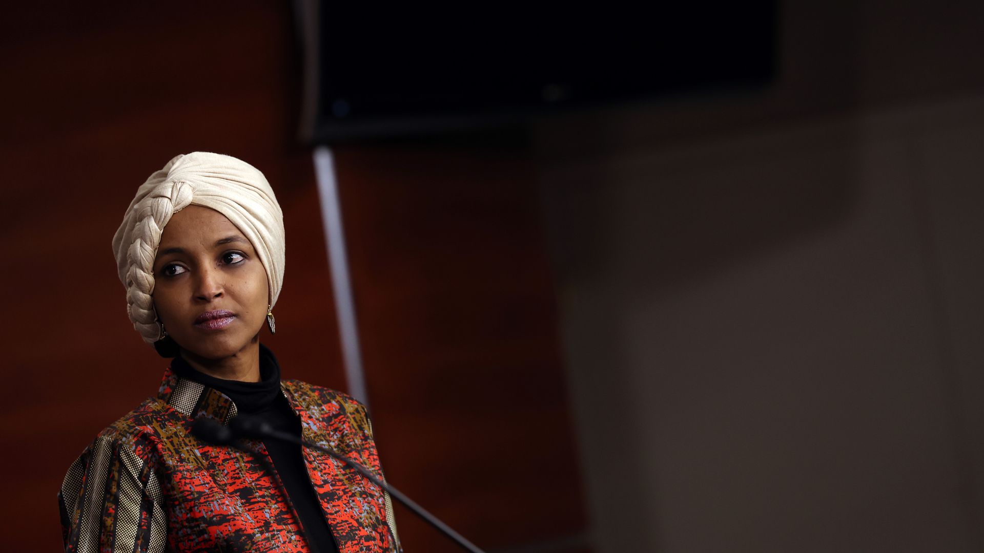 Rep. Ilhan Omar, wearing a white headscarf and a red, white, black and gold jacket over a blue sweater, speaks to reporters at a House TV studio.
