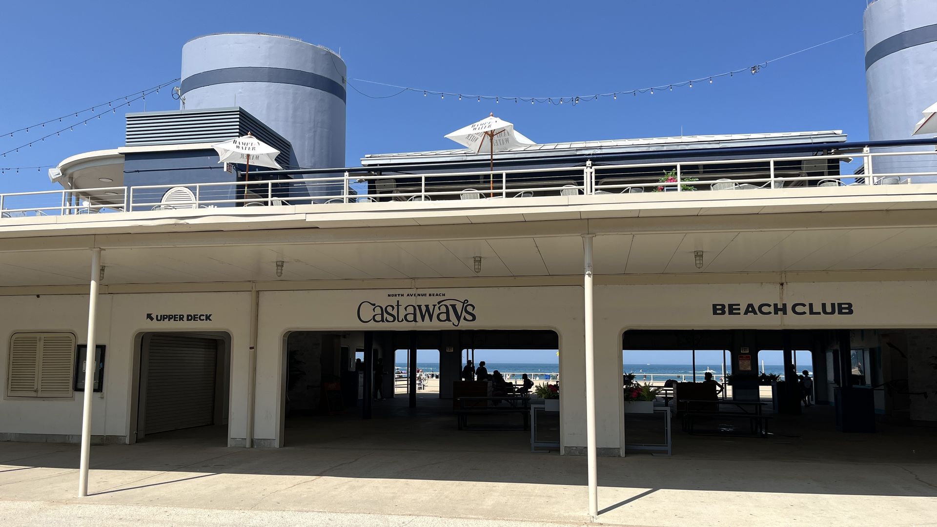 Two-floor beach club with patio with umbrellas and Lake Michigan in the distance.
